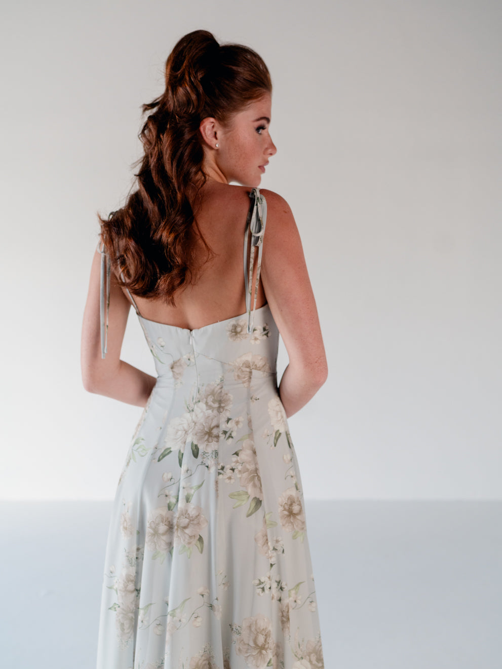 A woman shown from the back wearing a sleeveless, open-back A-line dress with thin tie straps and a soft floral print in muted beige and sage green on a pale blue-gray base, photographed against a plain light gray studio background. / A-line gown with double princess seam detail.