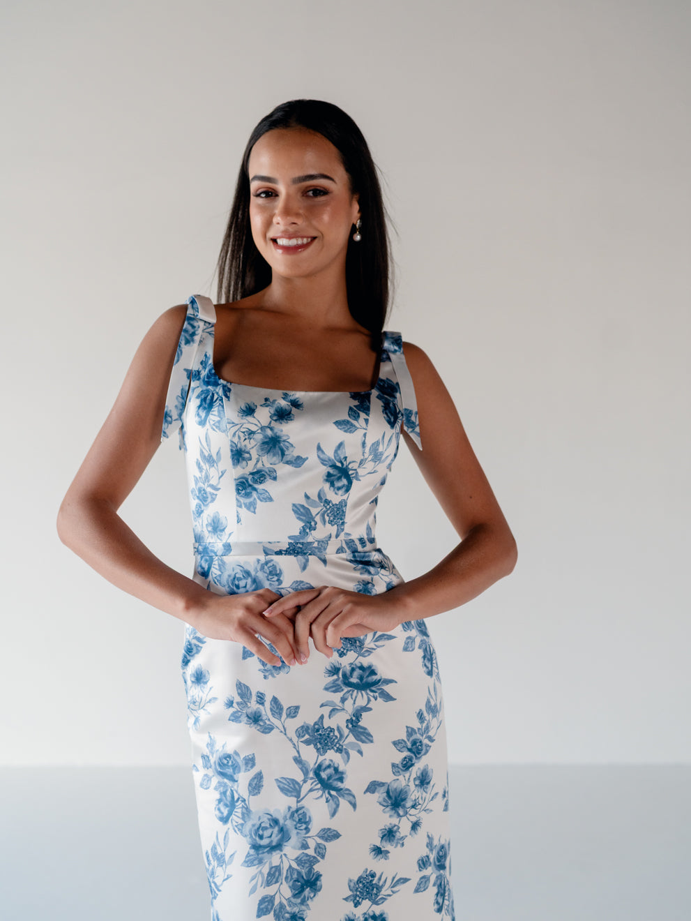 A woman modeling a sleeveless white dress with a blue floral toile pattern, featuring a square neckline and wide shoulder straps, set against a plain light gray background. / Slim satin gown with soft square neckline and bow detail at the shoulders.