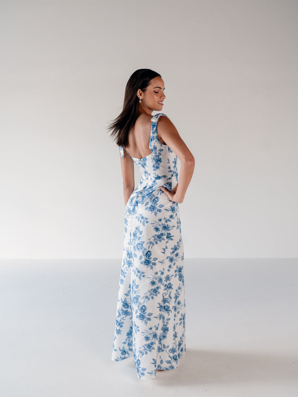 A woman modeling a sleeveless white maxi dress with a blue floral print, shown from the back in a studio setting with a plain light background. / Slim satin gown with soft square neckline and bow detail at the shoulders.
