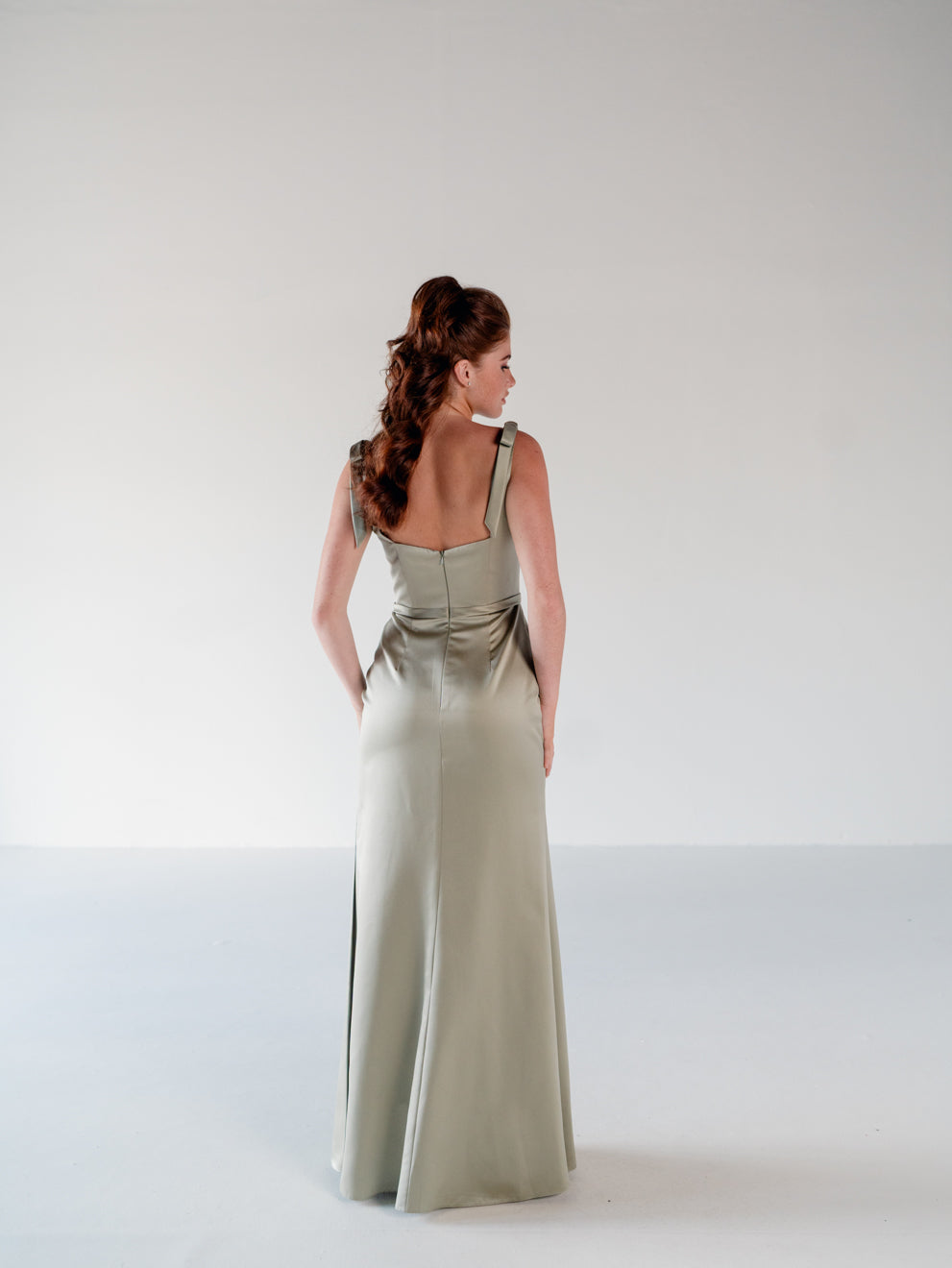 A woman stands with her back to the camera wearing a floor-length sage green satin gown with a low open back and thin shoulder straps, set against a plain light gray studio background. / Slim satin gown with soft square neckline and bow detail at the shoulders.