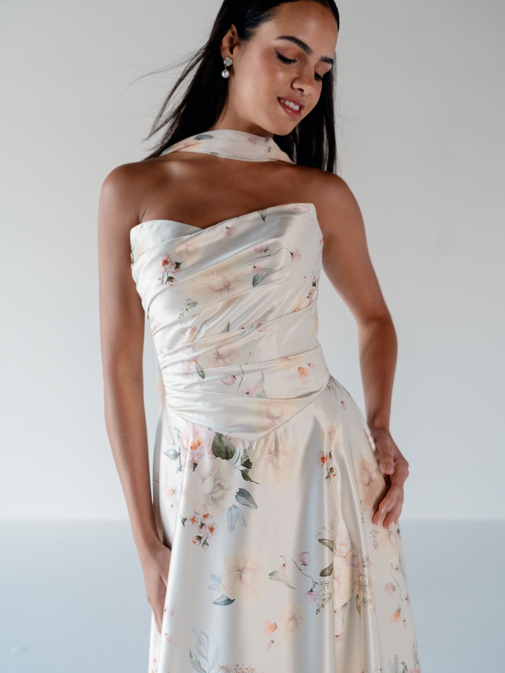 A woman modeling a strapless halter-neck midi dress with a draped bodice and a soft floral print in muted pink, green, and blue tones on a light cream background. / Basque waist, draped bodice A-line gown.