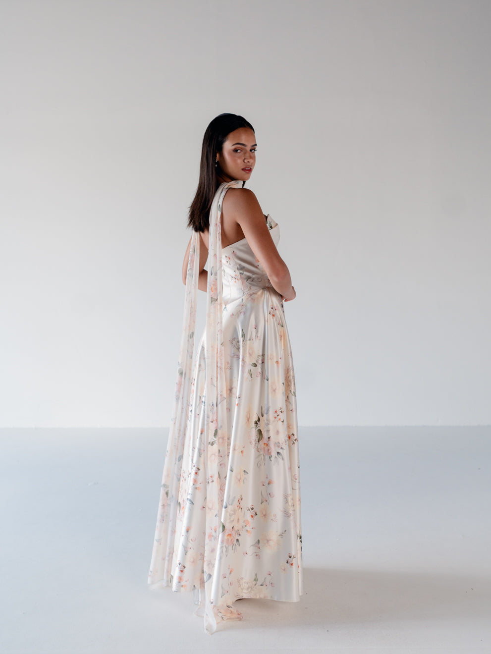 A woman stands in a studio wearing a sleeveless, floor-length cream dress with a soft floral print and sheer draped fabric over the back. / Basque waist, draped bodice A-line gown.