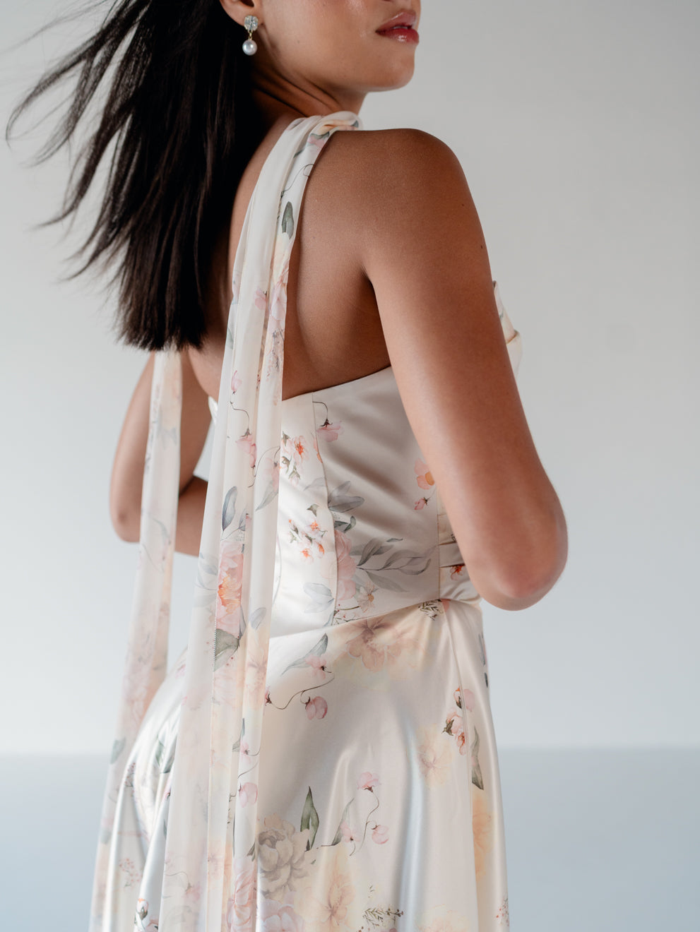 A woman shown from the back wearing a sleeveless satin dress with a sheer floral dupatta in soft blush, peach, and sage tones against a light gray background. / Basque waist, draped bodice A-line gown.