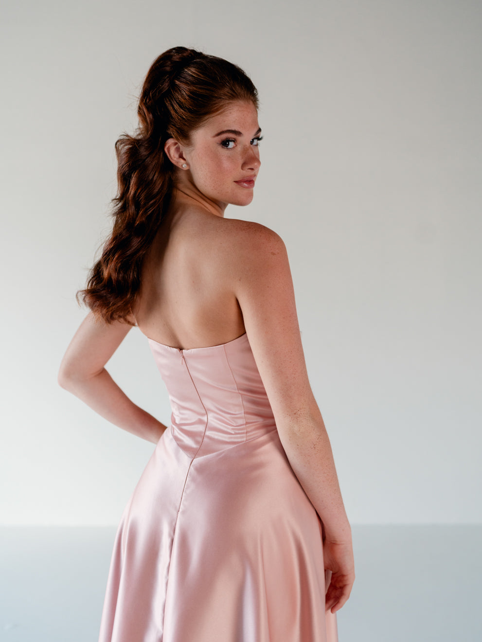 A woman wearing a strapless light pink satin gown, photographed from behind and slightly to the side against a plain light gray studio background. / Basque waist, draped bodice A-line gown.
