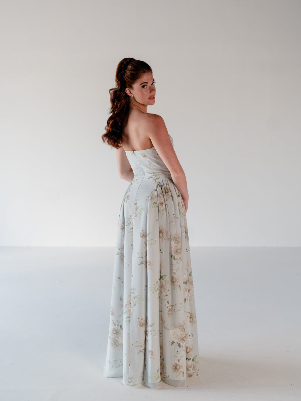 A woman stands in a studio wearing a strapless floor-length gown with a soft floral print. The dress has a light blue-gray base with beige and muted green floral motifs. / Draped neckline gown with gathered side detail.