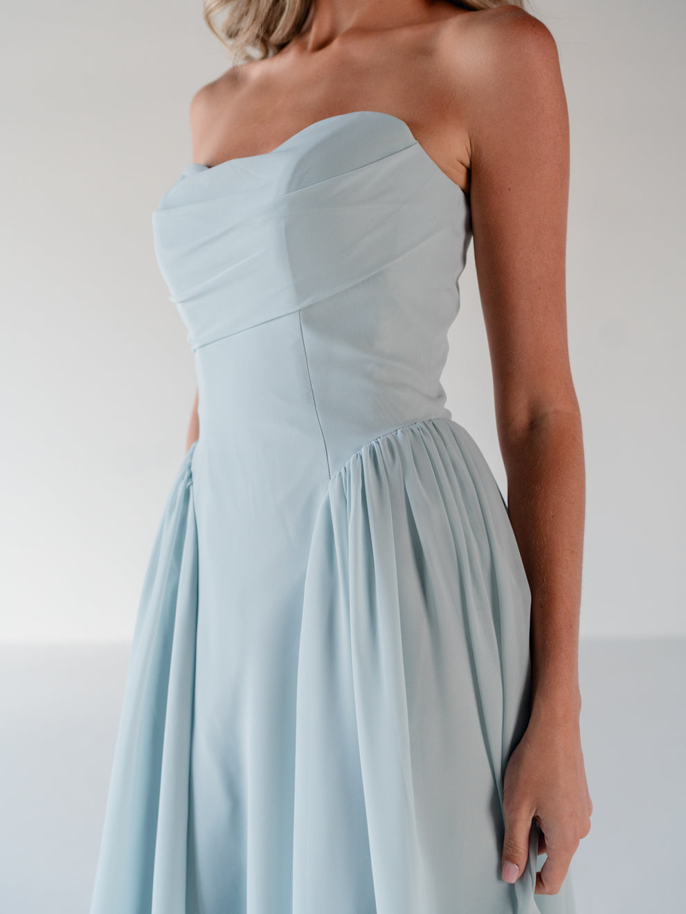 A woman wearing a strapless light blue dress with a structured sweetheart bodice and a flowing skirt, shown against a plain light gray studio background. / Draped neckline gown with gathered side detail.