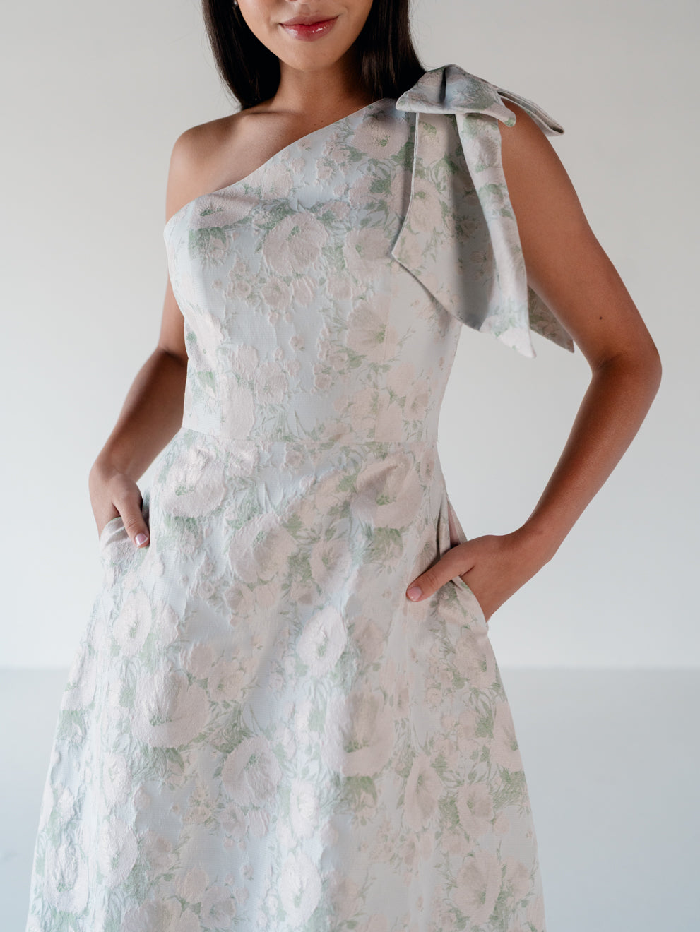 A woman wearing a one-shoulder white dress with a light green floral pattern and a matching fabric bow on the shoulder, photographed against a plain light background. / One shoulder gown with bow detail and ruffle at the skirt.
