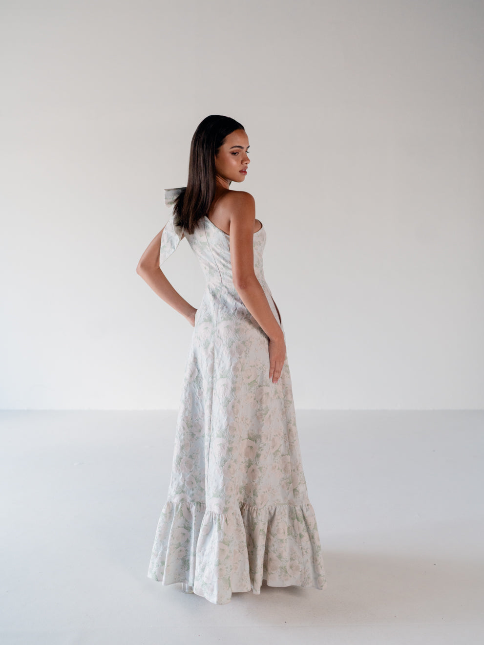 A woman modeling a strapless, floor-length floral dress with a ruffled hem, photographed in a minimalist studio setting with a white background. / One shoulder gown with bow detail and ruffle at the skirt.