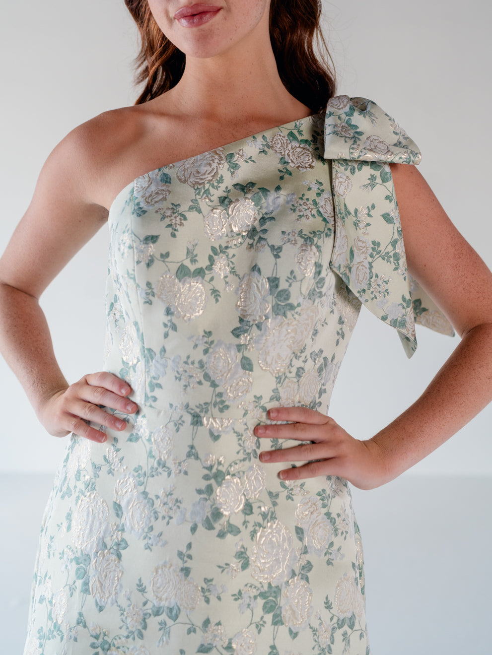 A woman wearing a one-shoulder formal dress with a large bow detail on the shoulder, featuring a light beige base with a green floral pattern and metallic rose-gold floral highlights. / One shoulder gown with bow detail and ruffle at the skirt.