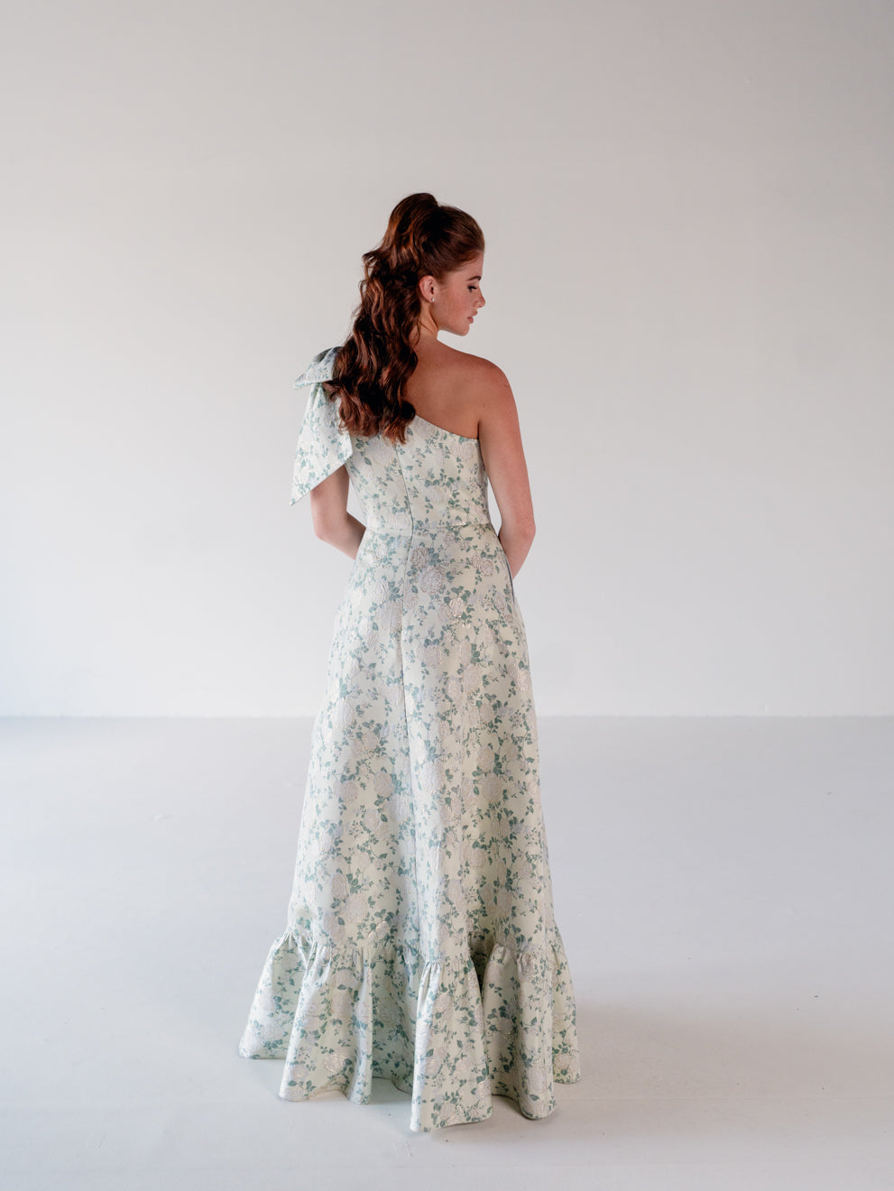 A woman stands with her back to the camera wearing a one-shoulder maxi dress with a delicate green floral print on a cream base, set against a plain light gray studio background. / One shoulder gown with bow detail and ruffle at the skirt.