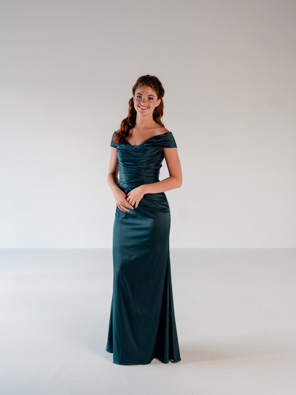 A woman modeling a floor-length off-the-shoulder gown in a deep teal satin-like fabric, standing against a plain light gray studio background. / Off the shoulder, stretch matte charmeuse gown with asymmetrical draping.