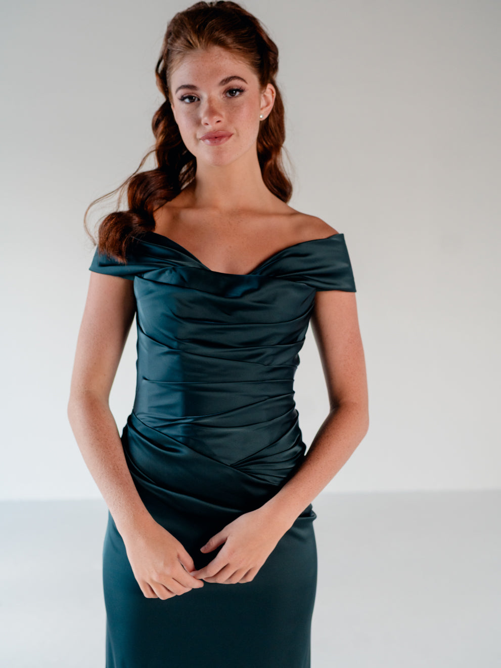 A woman modeling an off-the-shoulder dark teal satin dress with ruched detailing on a light gray studio background. / Off the shoulder, stretch matte charmeuse gown with asymmetrical draping.