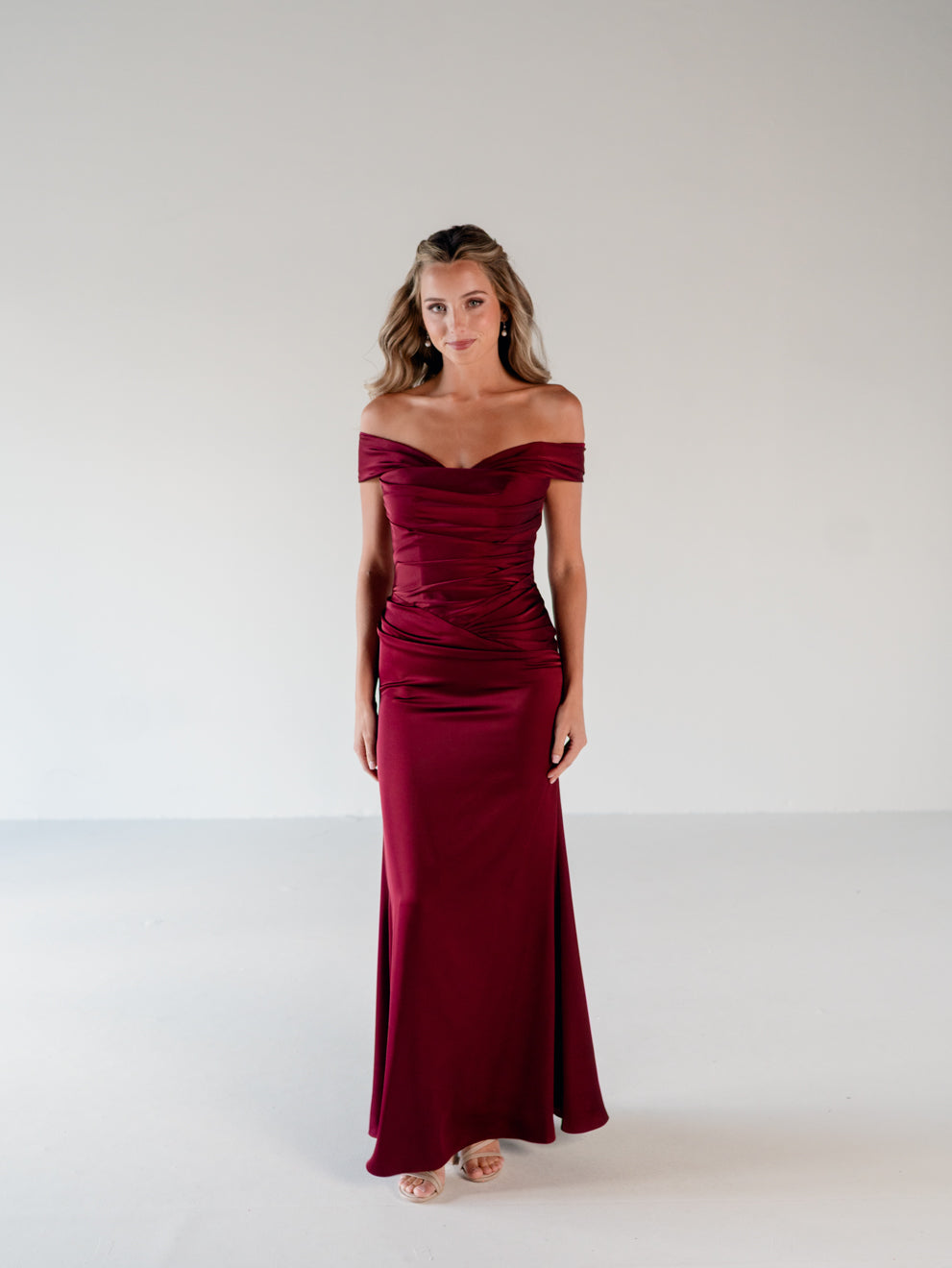 A woman modeling a floor-length off-the-shoulder gown in deep burgundy with a ruched bodice and satin-like sheen, standing against a plain light gray studio background. / Off the shoulder, stretch matte charmeuse gown with asymmetrical draping.