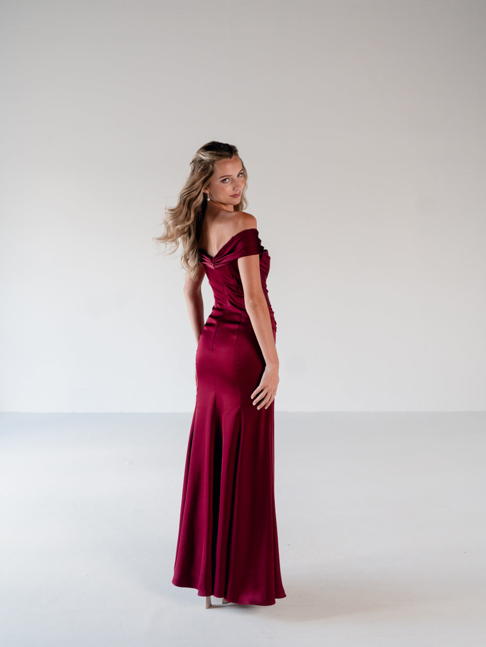 A woman modeling a floor-length off-the-shoulder evening gown in deep burgundy satin, standing against a plain light gray studio background. / Off the shoulder, stretch matte charmeuse gown with asymmetrical draping.