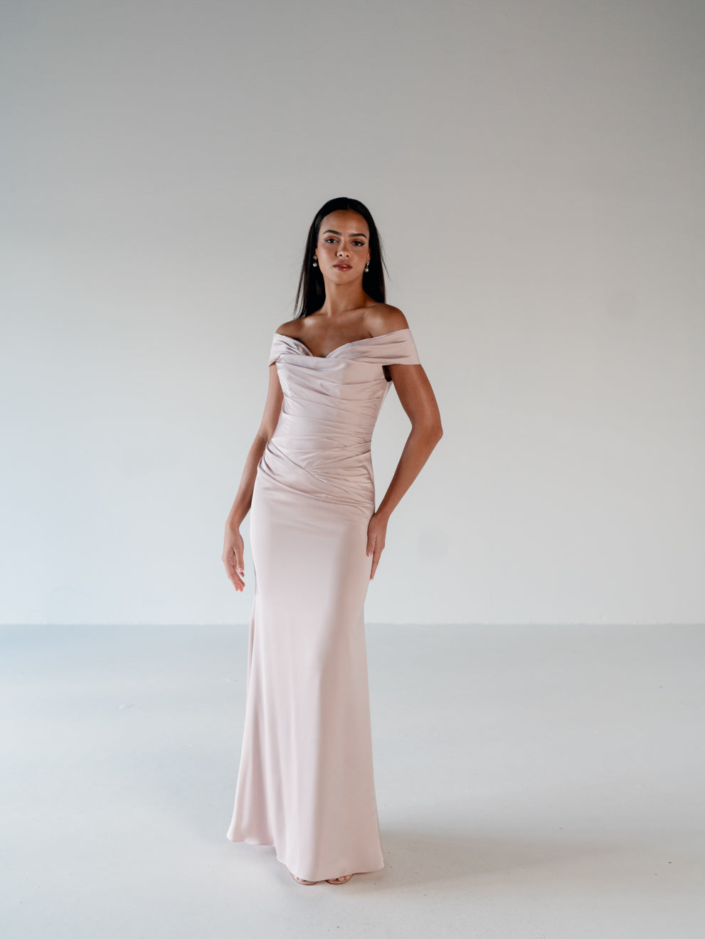 A woman modeling a floor-length off-the-shoulder gown in a pale blush pink color, standing against a neutral light gray studio background. / Off the shoulder, stretch matte charmeuse gown with asymmetrical draping.