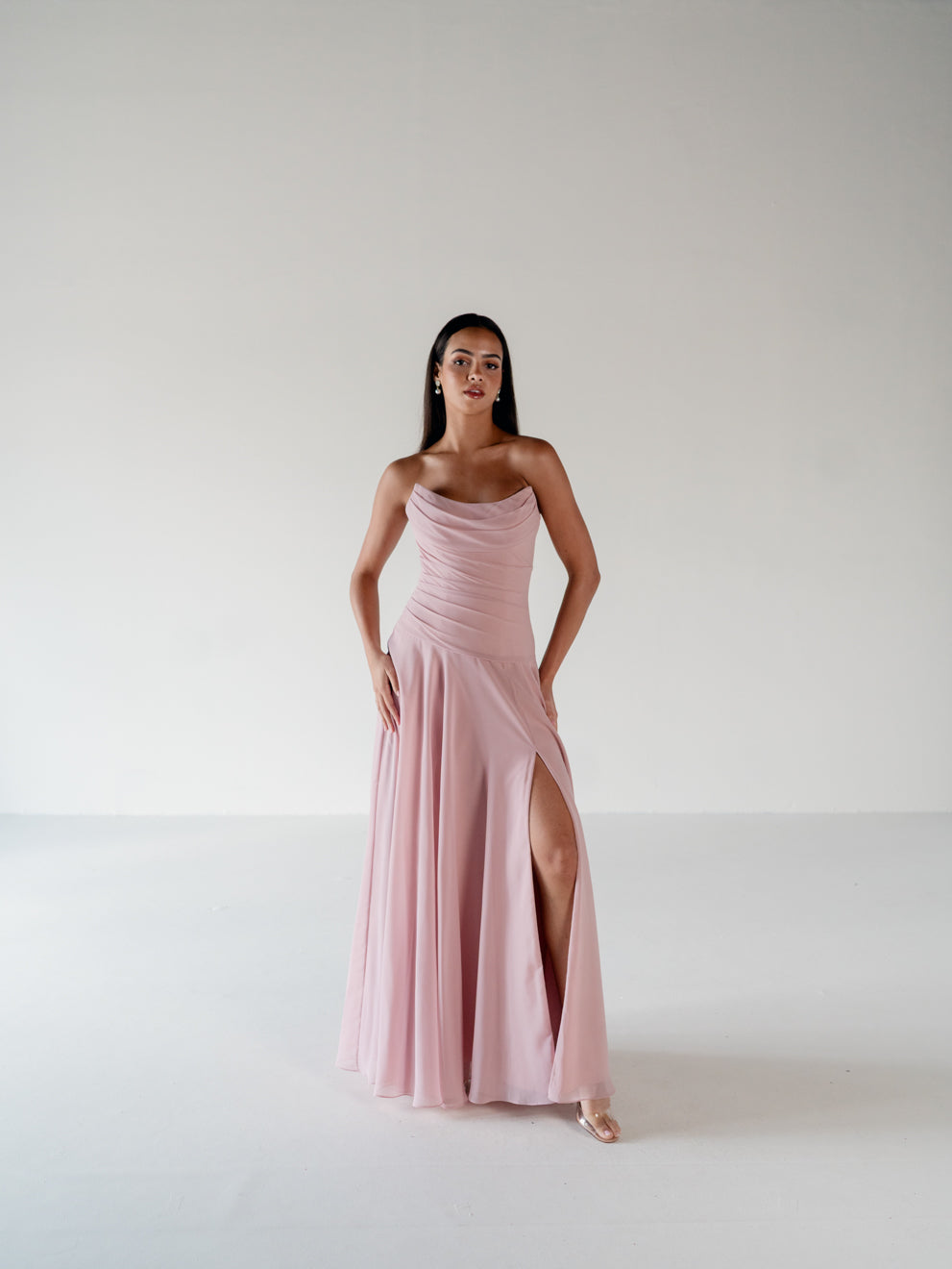 A woman modeling a floor-length, strapless dusty pink gown with a draped bodice, flowing skirt, and high front slit, standing against a plain light gray studio background. / Strapless draped bodice with asymmetrical placed skirt and lace up back.