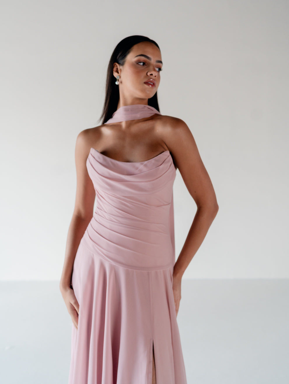 A woman modeling a dusty pink halter-neck midi dress with draped ruching on the bodice and a high slit, set against a plain light gray studio background. / Strapless draped bodice with asymmetrical placed skirt and lace up back.