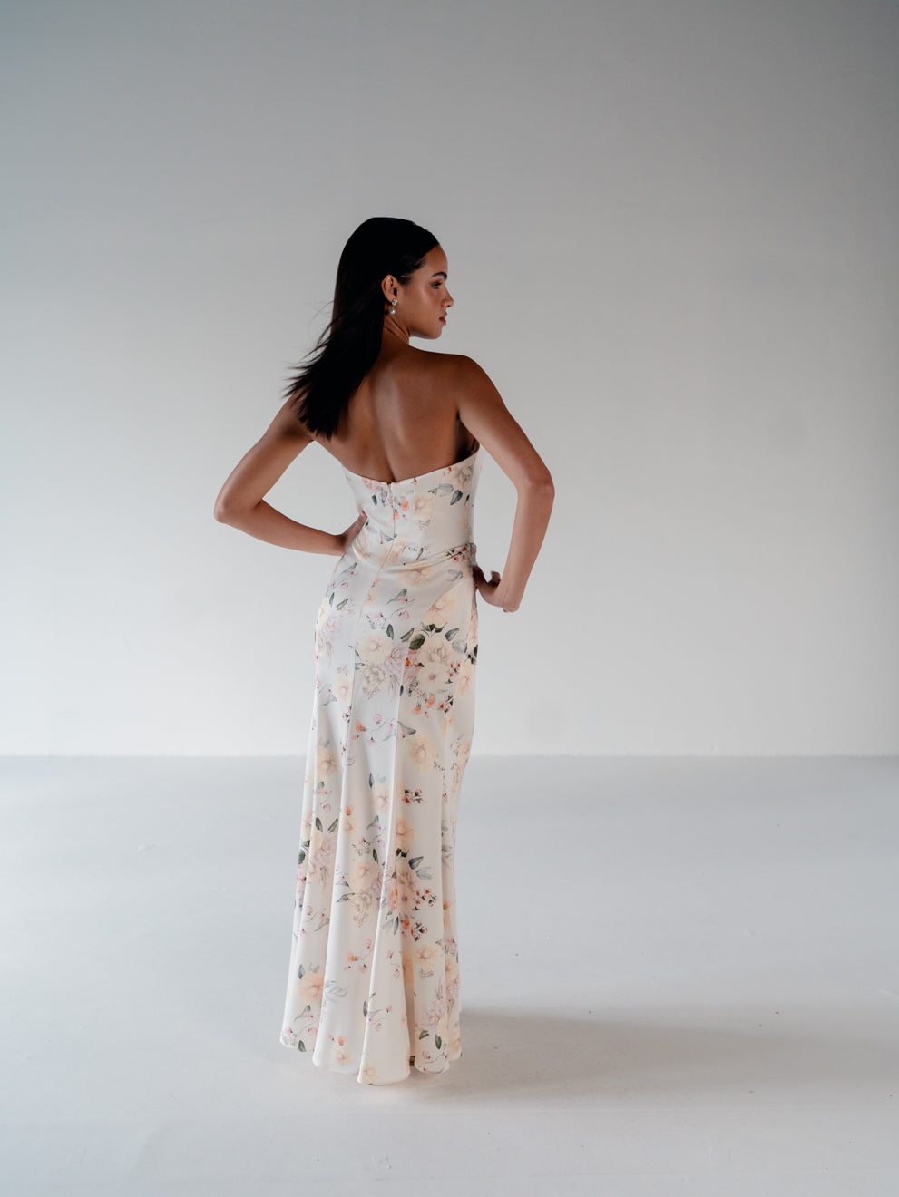 A woman stands with her back to the camera wearing a strapless, floor-length floral gown in a light cream color with pink, green, and blue floral details, set against a plain light gray studio background. / Scoop neckline fitted bodice with draped skirt.
