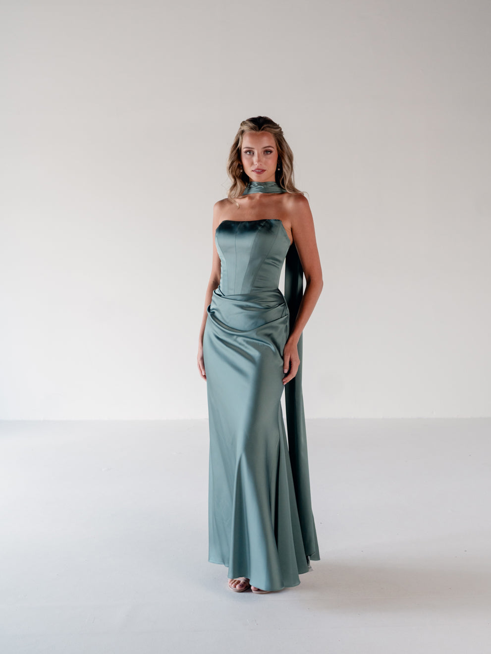 A woman modeling a floor-length satin gown in a dusty sage green color, featuring a halter-style neckline with a draped side detail, set against a plain light gray studio background. / Scoop neckline fitted bodice with draped skirt.