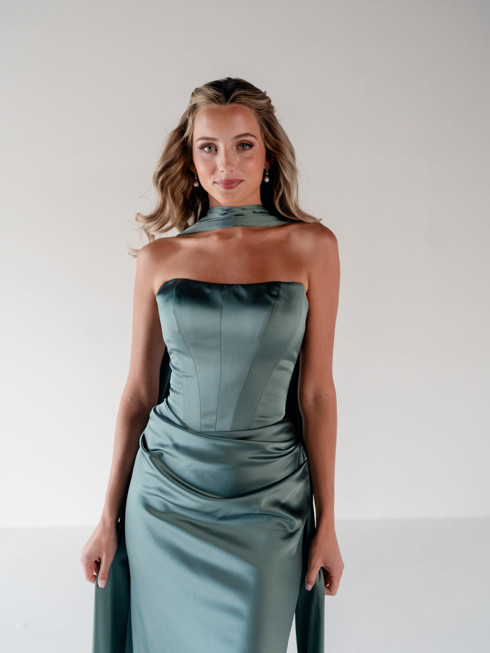 A woman modeling a strapless satin gown in a muted sage green color, featuring a structured corset bodice and draped side details, set against a plain light gray studio background. / Scoop neckline fitted bodice with draped skirt.