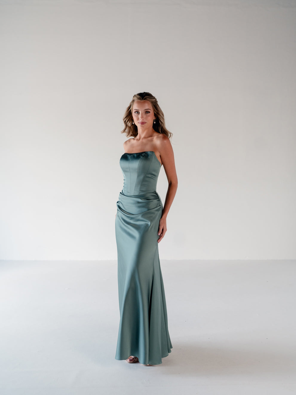 A woman modeling a strapless satin floor-length gown in a dusty sage green color, standing against a plain white studio background. / Scoop neckline fitted bodice with draped skirt.