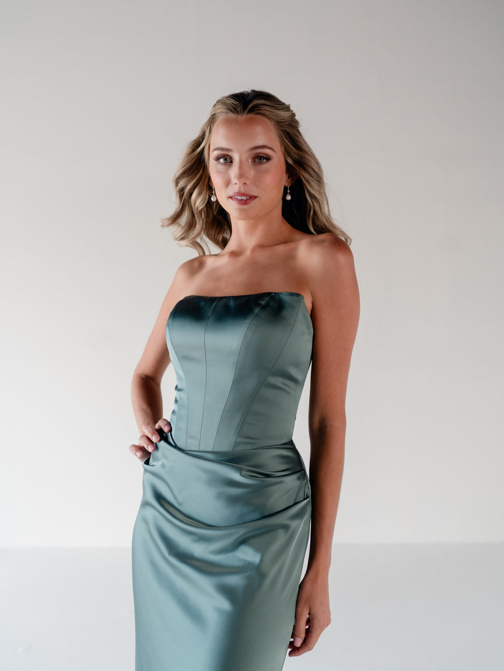 A woman modeling a strapless satin midi dress in a dusty sage green color, standing against a neutral light gray studio background. / Scoop neckline fitted bodice with draped skirt.