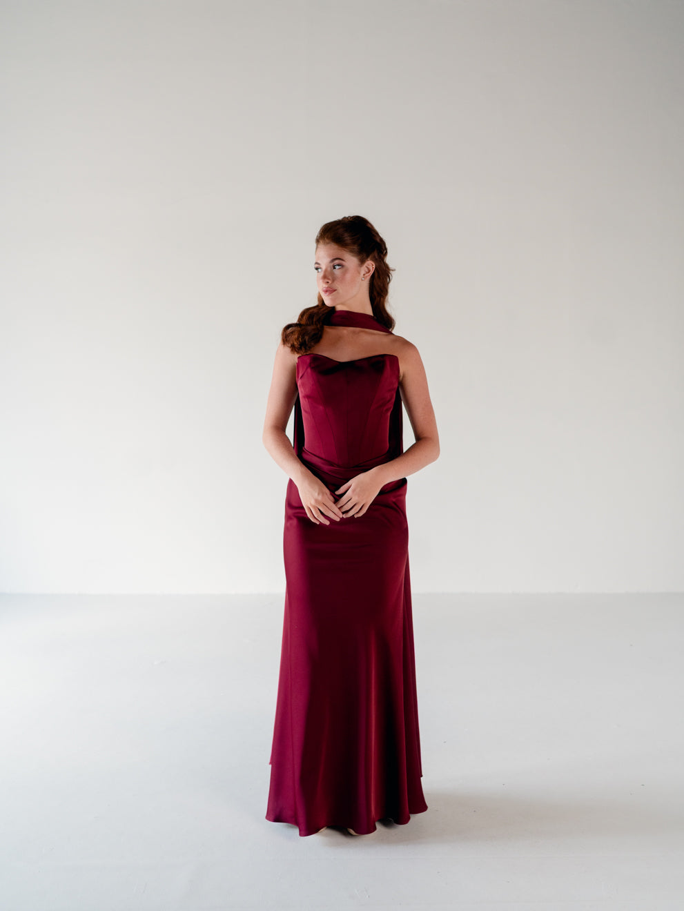 A woman modeling a floor-length burgundy evening gown with a halter-style neckline and draped ruffle detail, standing against a plain light gray studio background. / Scoop neckline fitted bodice with draped skirt.
