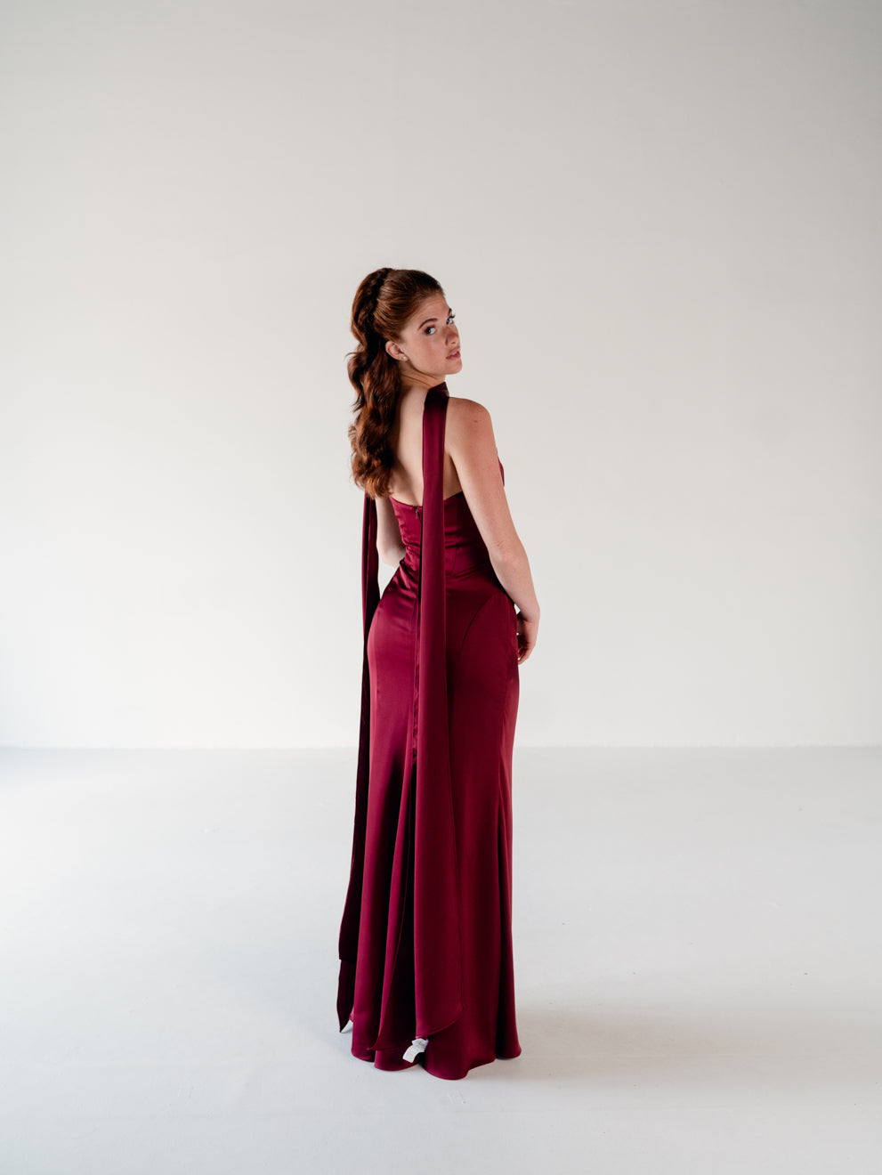 A woman modeling a floor-length burgundy evening gown with a draped, asymmetrical back detail, photographed in a minimalist white studio setting. / Scoop neckline fitted bodice with draped skirt.