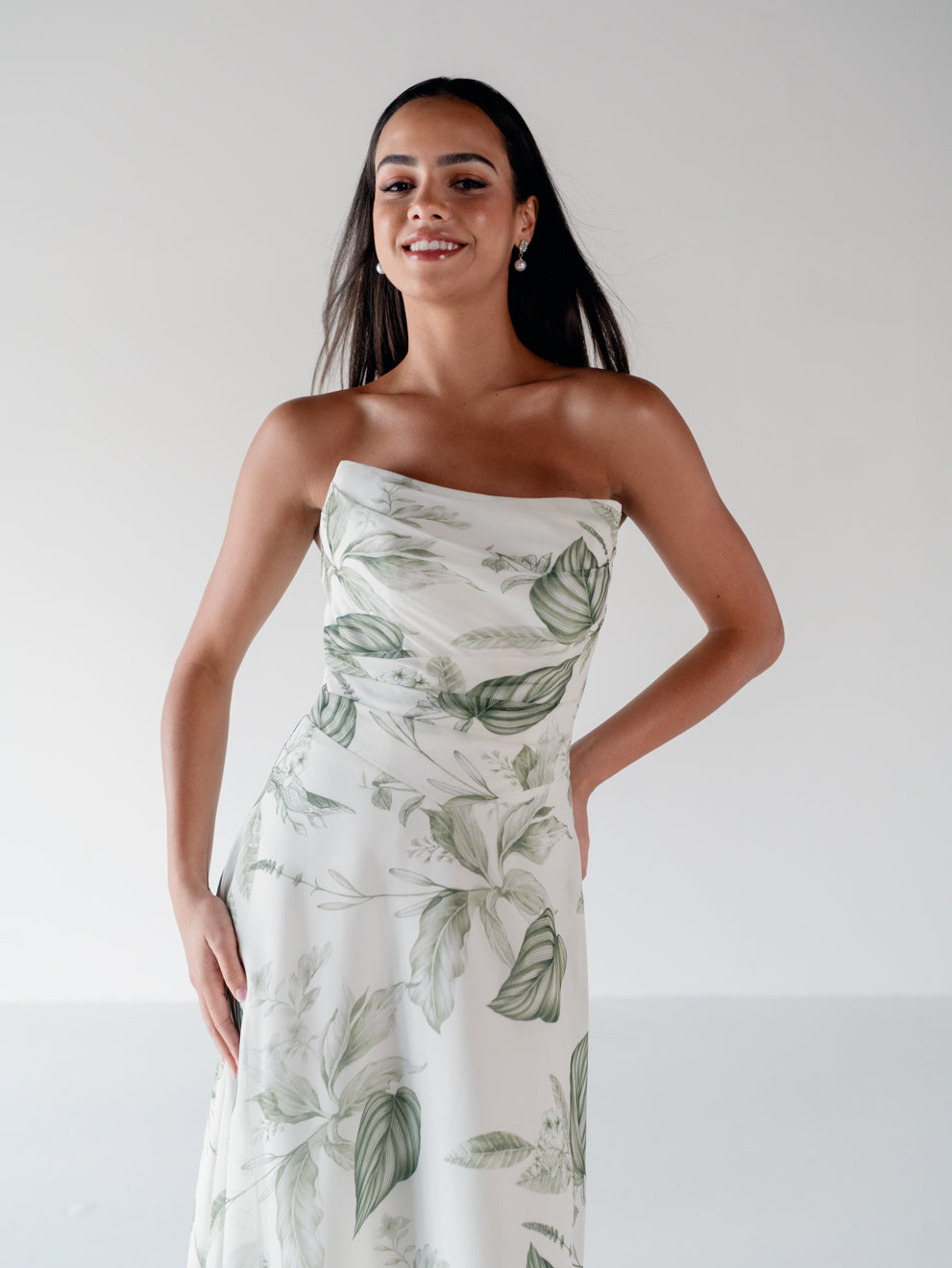 A woman modeling a strapless white dress with a green tropical leaf print, set against a plain light gray studio background. / Draped bodice with A-line skirt and lace up back.