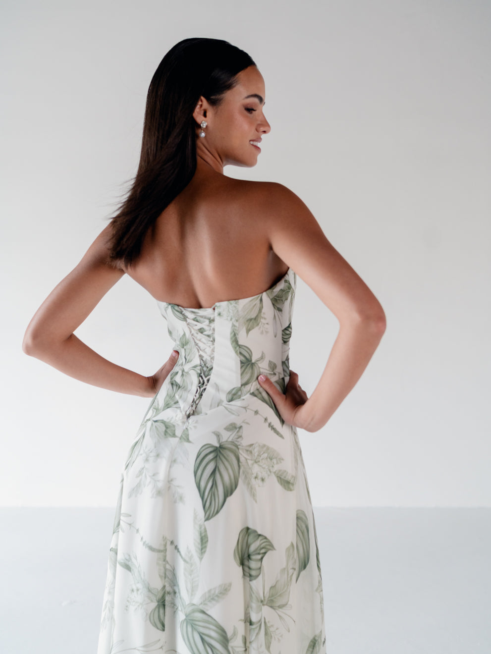 A woman modeling a strapless white dress with a green tropical leaf print, shown from the back against a plain light gray studio background. / Draped bodice with A-line skirt and lace up back.