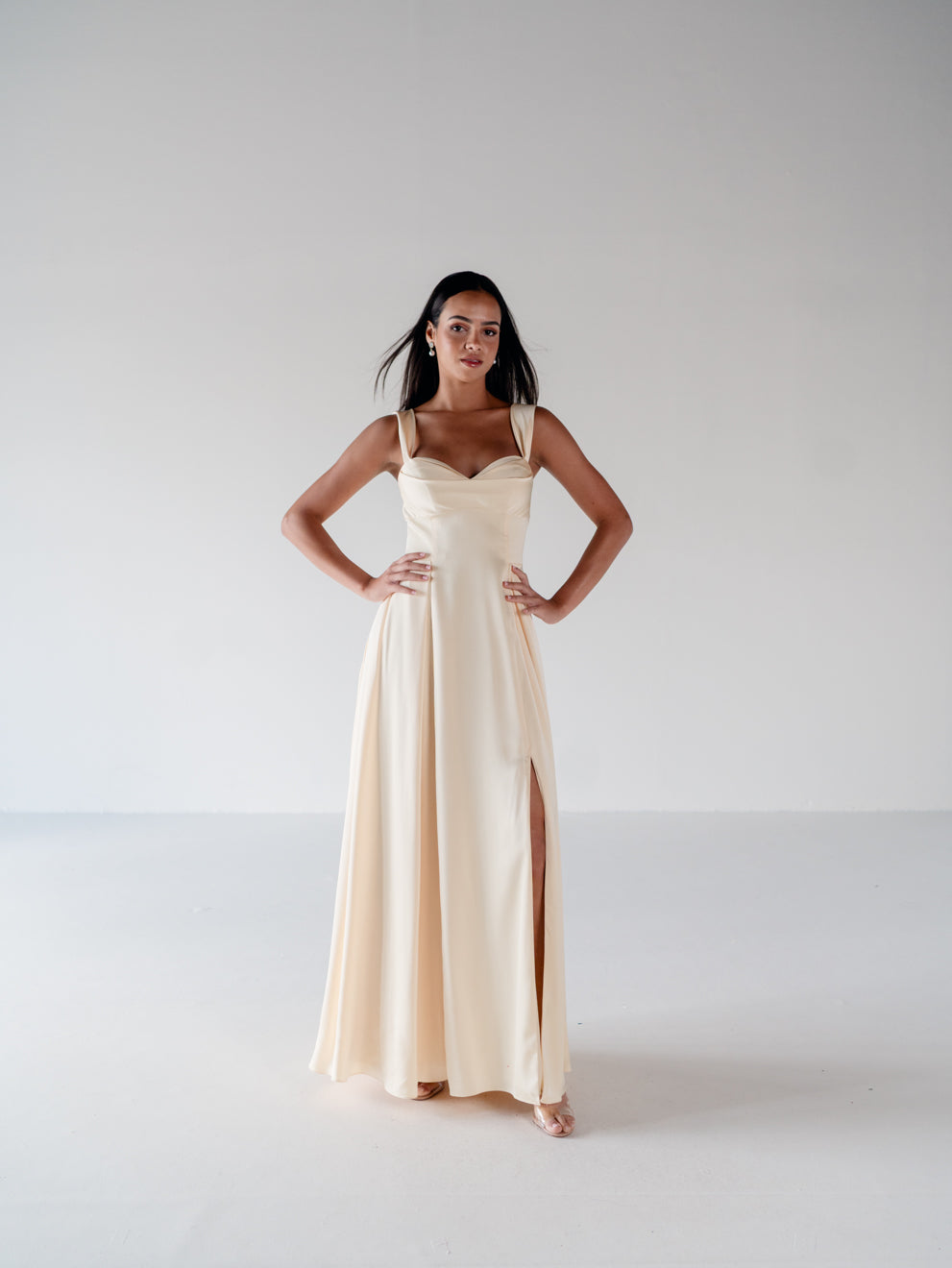 A woman modeling a floor-length cream satin gown with a sweetheart neckline, thin shoulder straps, and a high side slit, standing against a seamless light gray studio background. / Draped neckline A-line gown with faux overskirt and slit detail.