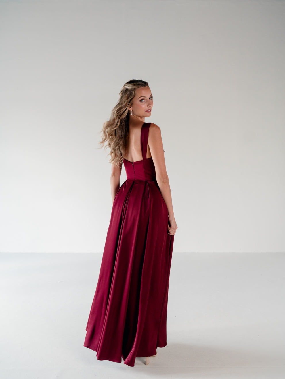 A woman modeling a floor-length burgundy satin gown from the back, standing against a seamless light gray studio background. / Draped neckline A-line gown with faux overskirt and slit detail.