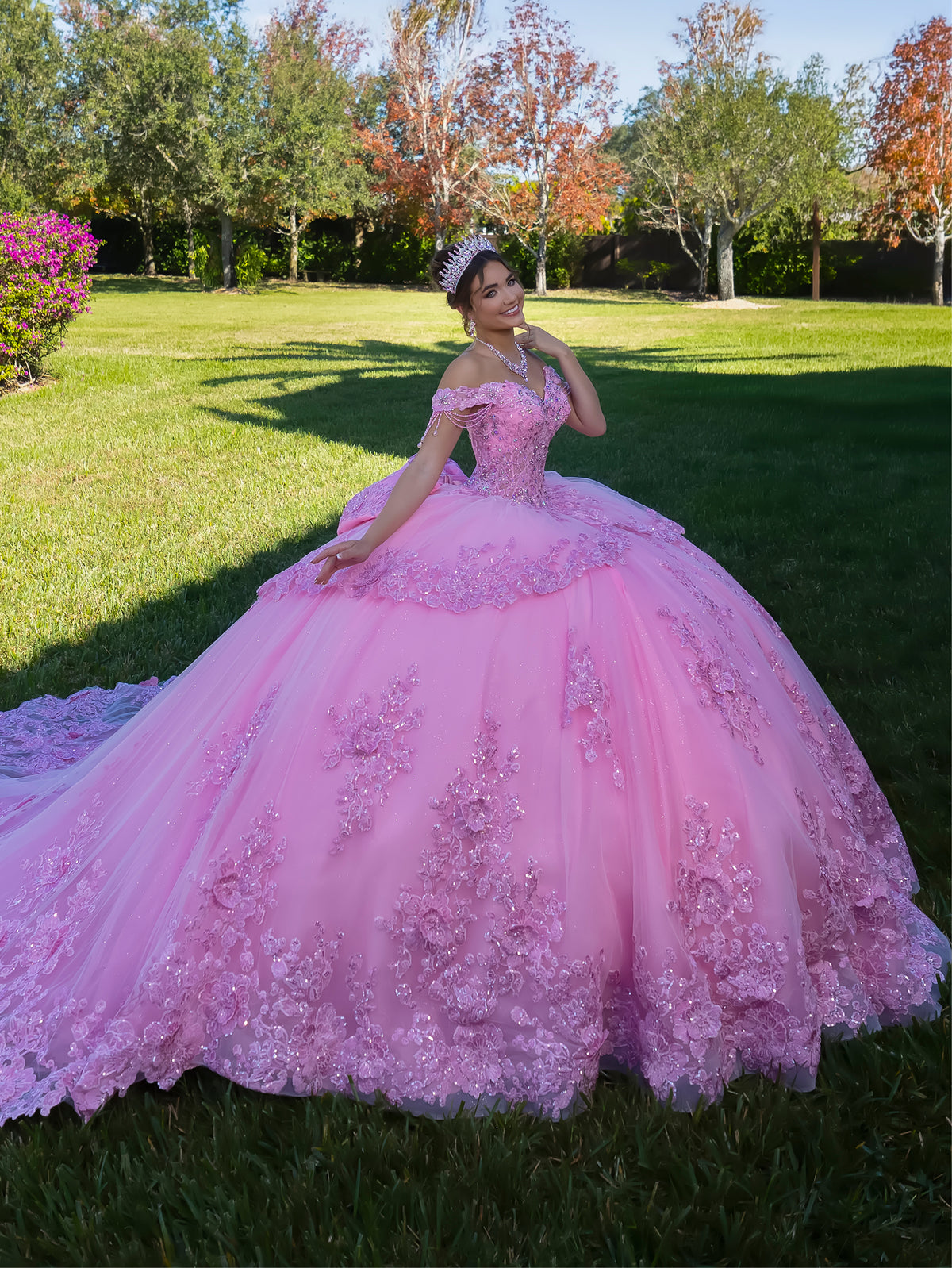 Woman in a pink quinceañera dress standing outdoors on grass with trees in the background / This ball gown features a richly detailed bodice with floral appliqué and off-the-shoulder straps. The skirt is extremely full and layered with cascading floral accents, creating a dramatic, fairytale look.
