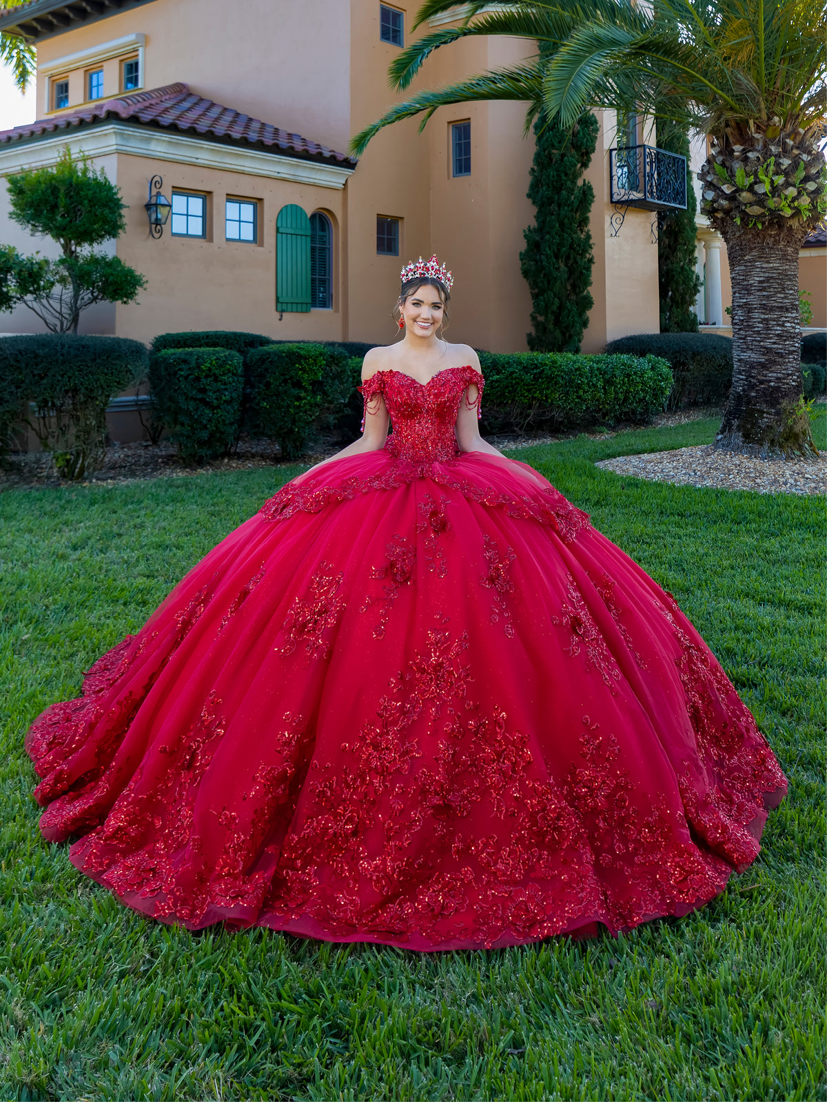 Woman in a red ball gown standing in front of a building with greenery / This ball gown features a richly detailed bodice with floral appliqué and off-the-shoulder straps. The skirt is extremely full and layered with cascading floral accents, creating a dramatic, fairytale look.