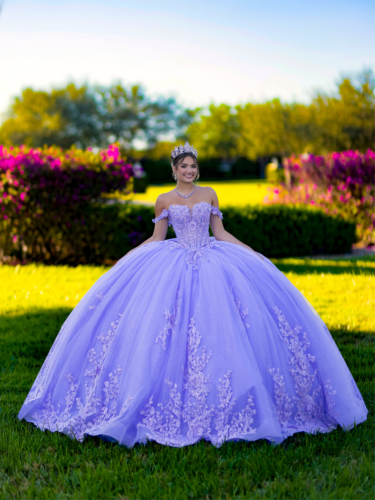 Woman in a purple ball gown standing in a garden with colorful flowers and greenery. / This ball gown features an ornate bodice with floral appliqué and off-the-shoulder straps. The skirt is extremely full of layered tulle and cascading floral details, creating a dramatic, fairytale silhouette.