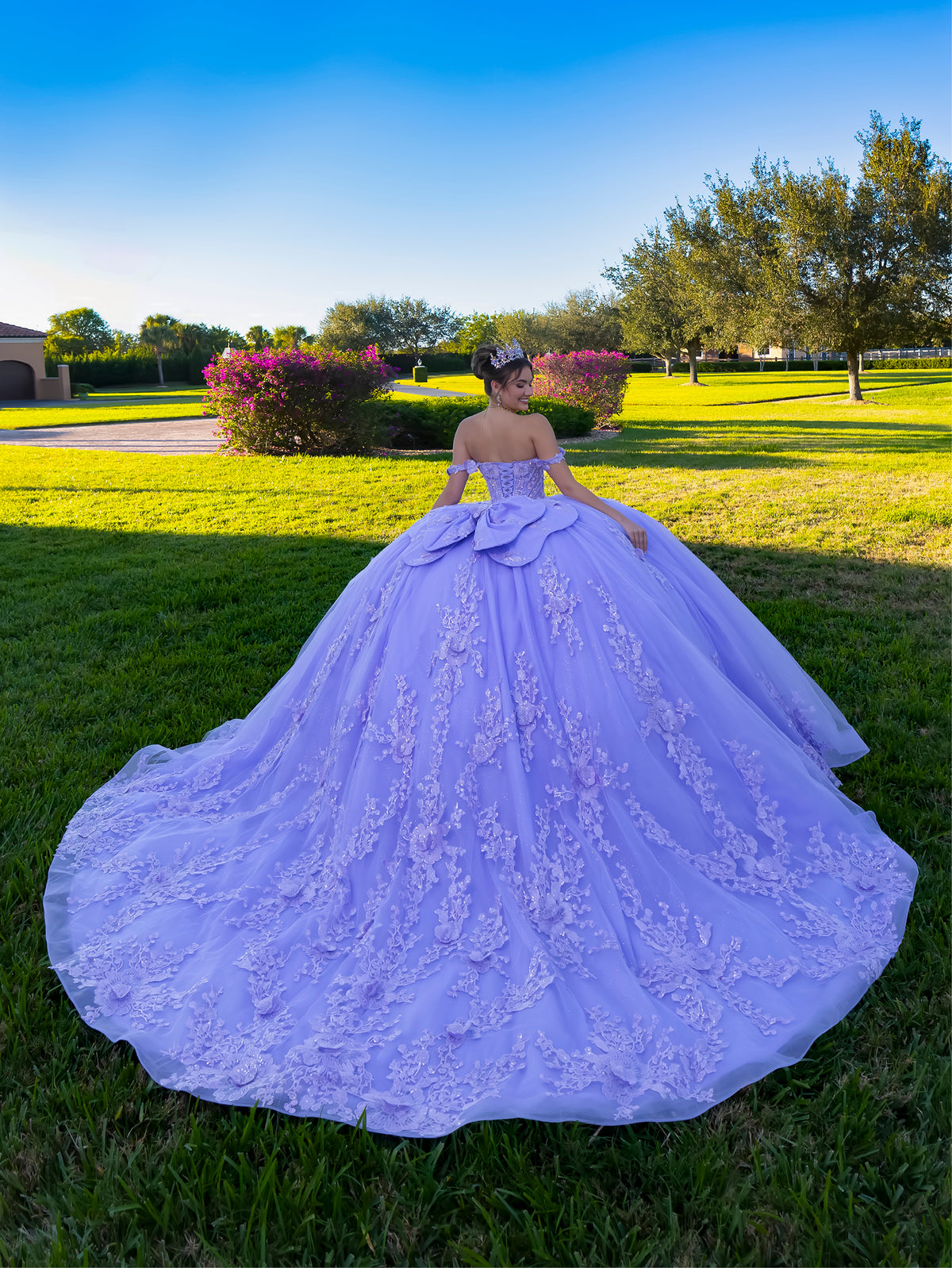 A woman stands outdoors on a grassy lawn wearing a voluminous lavender ball gown with floral lace applique and a large bow at the back of the neckline. Trees, shrubs, and a blue sky are visible in the background. / This ball gown features an ornate bodice with floral appliqué and off-the-shoulder straps. The skirt is extremely full of layered tulle and cascading floral details, creating a dramatic, fairytale silhouette.