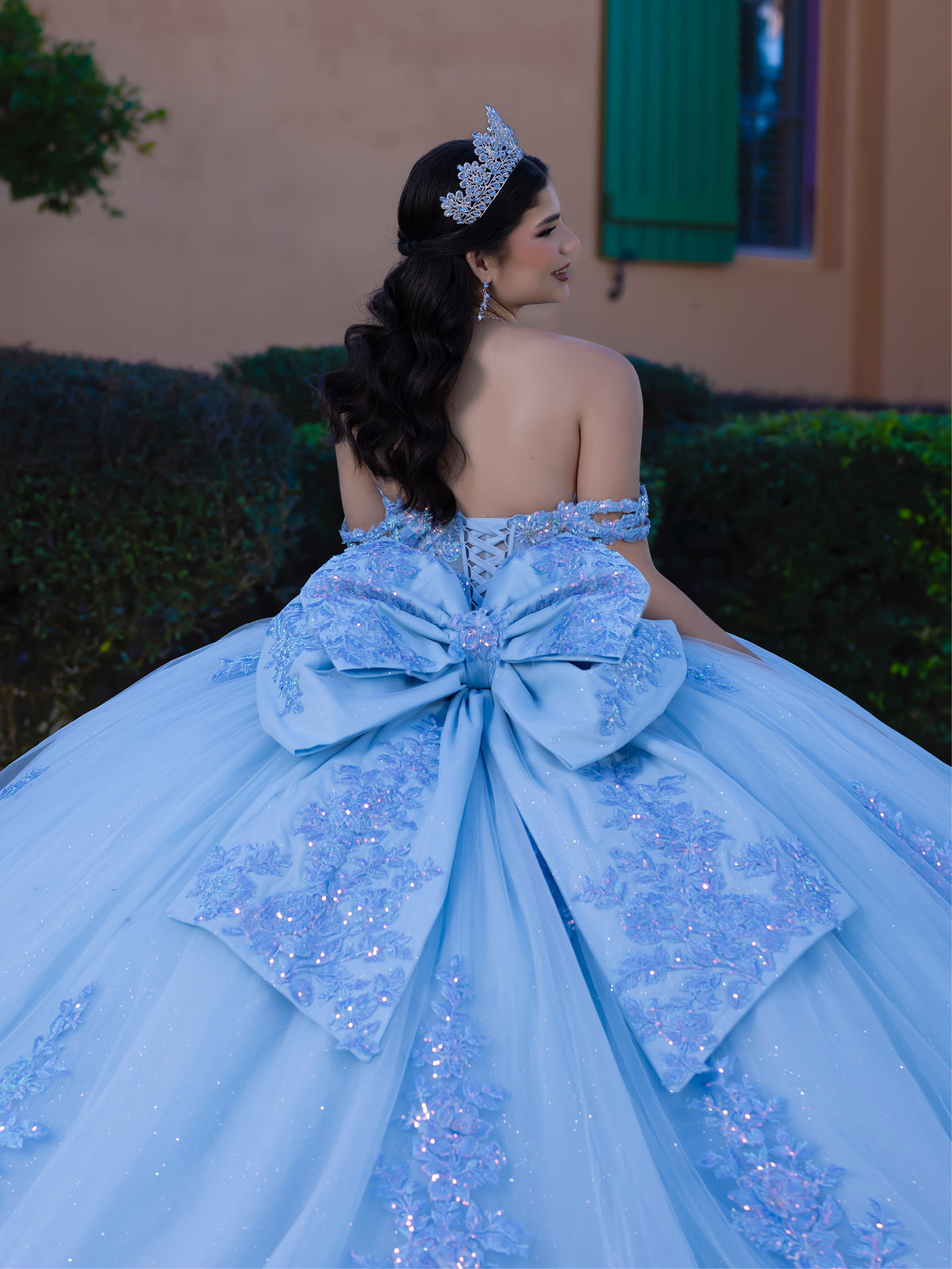 Woman in a blue quinceañera dress with a large bow, standing outdoors. / This ball gown features a detailed floral-appliqué bodice with soft off-the-shoulder straps. The full layered skirt is adorned with matching floral designs and flows into a dramatic, elegant train.