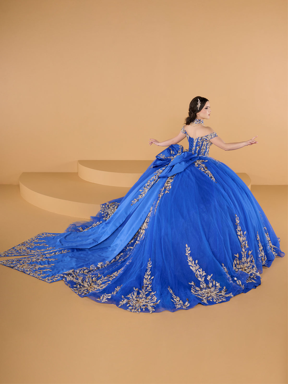 This ball gown features an ornate bodice with detailed embroidery and off-the-shoulder straps. The full tulle skirt is embellished with matching floral designs and finished with a dramatic bow and sweeping train.