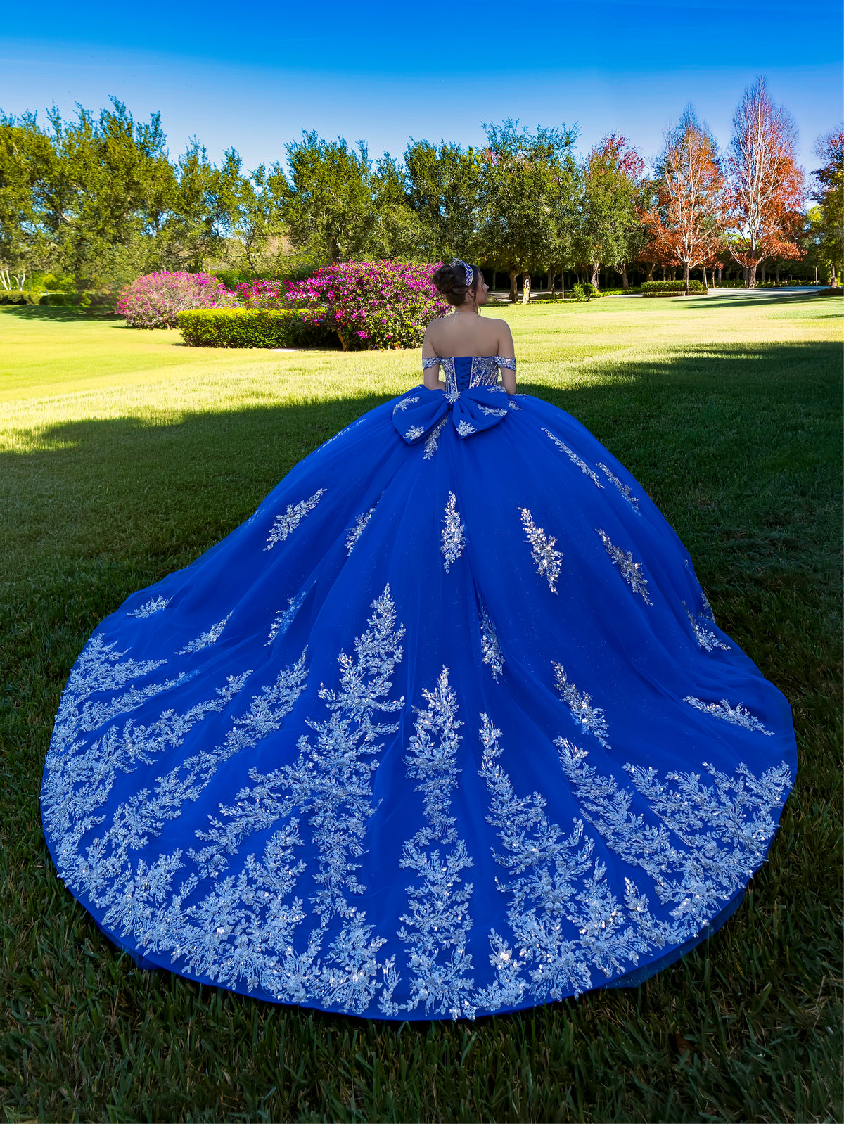 This ball gown features a detailed embroidered bodice and off-the-shoulder straps. The full, dramatic skirt is adorned with shimmering floral patterns and finishes with a large bow and flowing train.