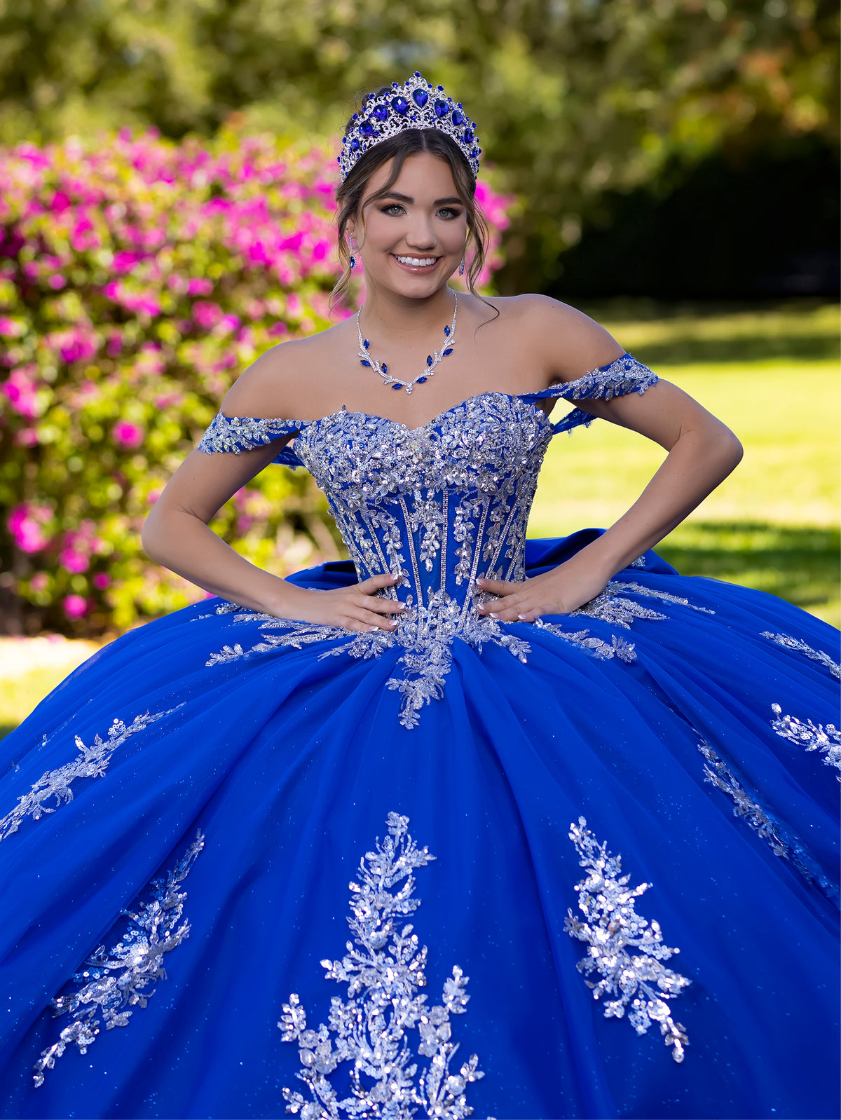 This ball gown features a detailed embroidered bodice and off-the-shoulder straps. The full, dramatic skirt is adorned with shimmering floral patterns and finishes with a large bow and flowing train.
