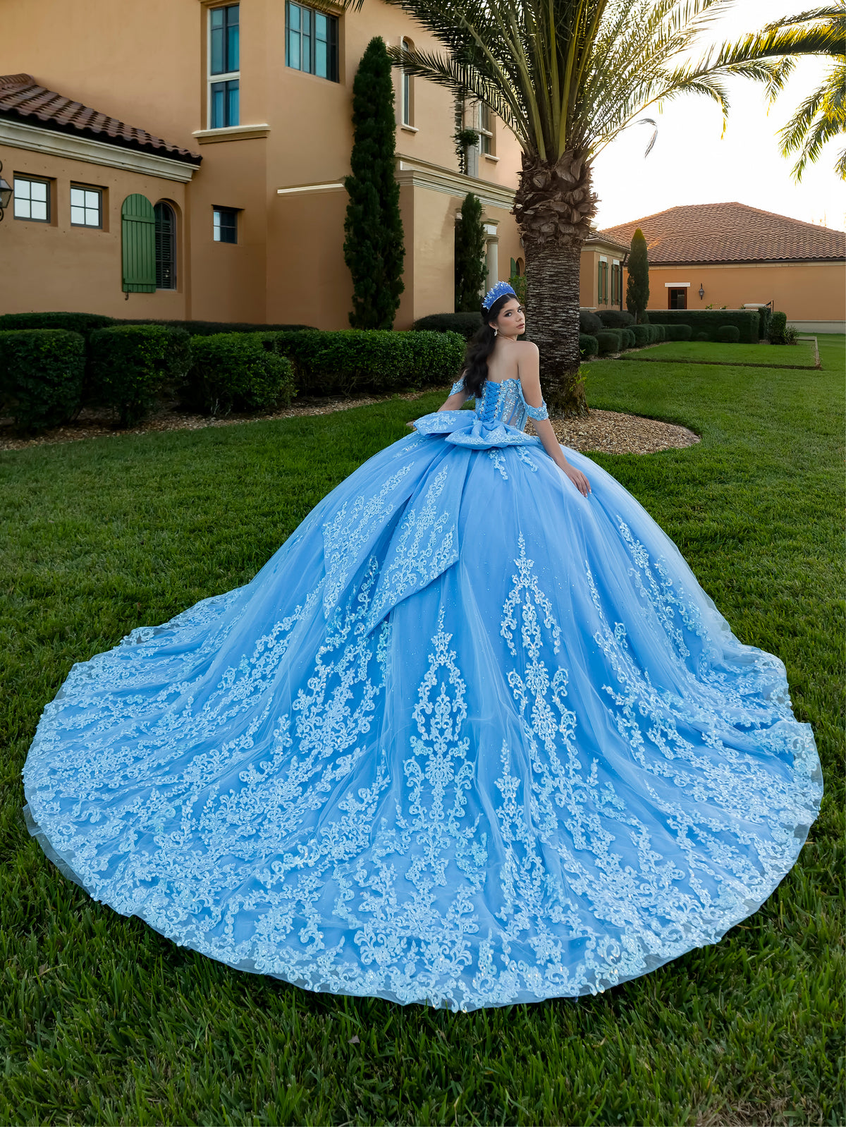 This ball gown features a richly embroidered bodice with off-the-shoulder straps. The full, voluminous skirt is decorated with shimmering floral detailing and flows into an elegant train accented with a bow at the back.