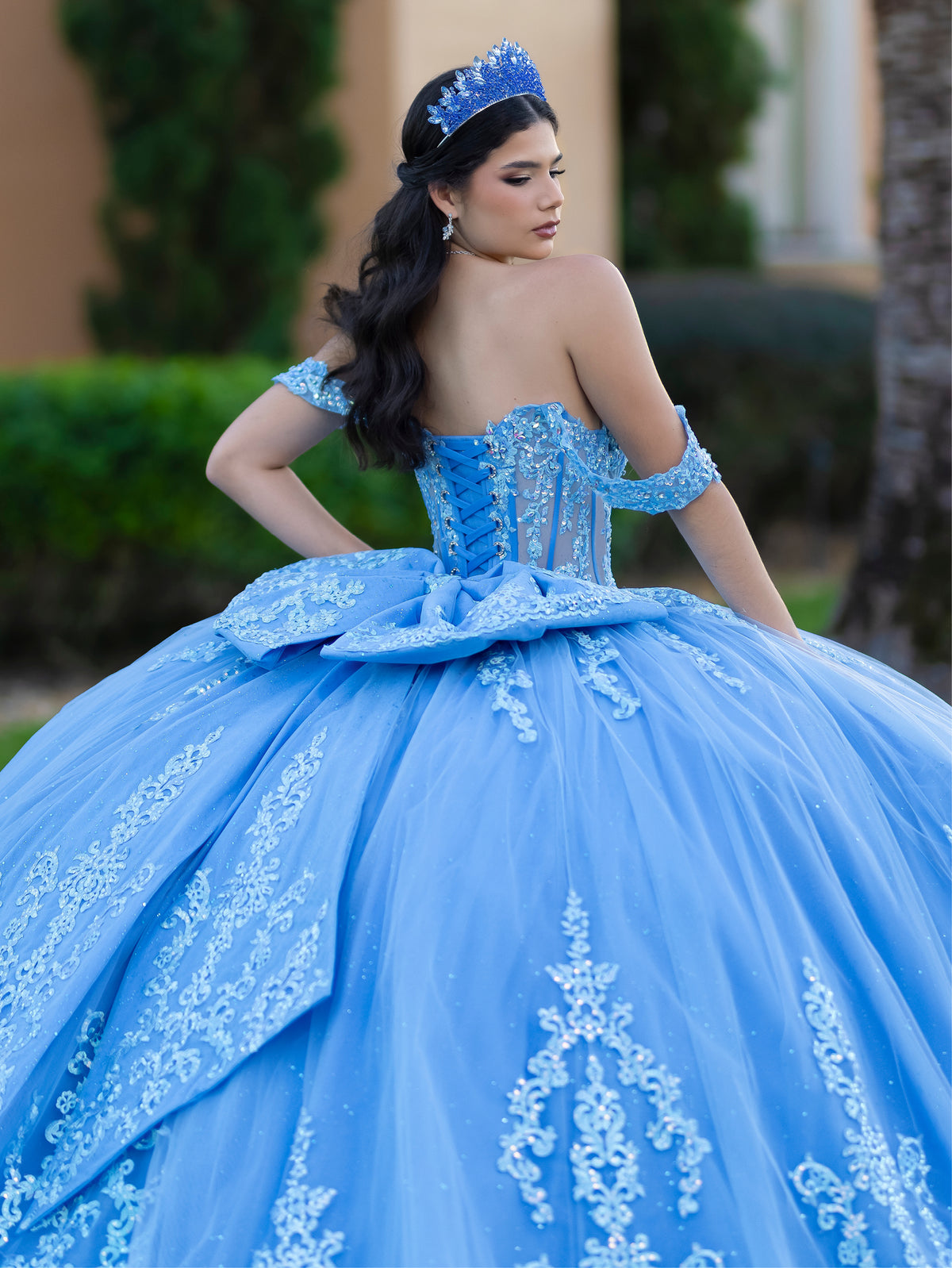 This ball gown features a richly embroidered bodice with off-the-shoulder straps. The full, voluminous skirt is decorated with shimmering floral detailing and flows into an elegant train accented with a bow at the back.