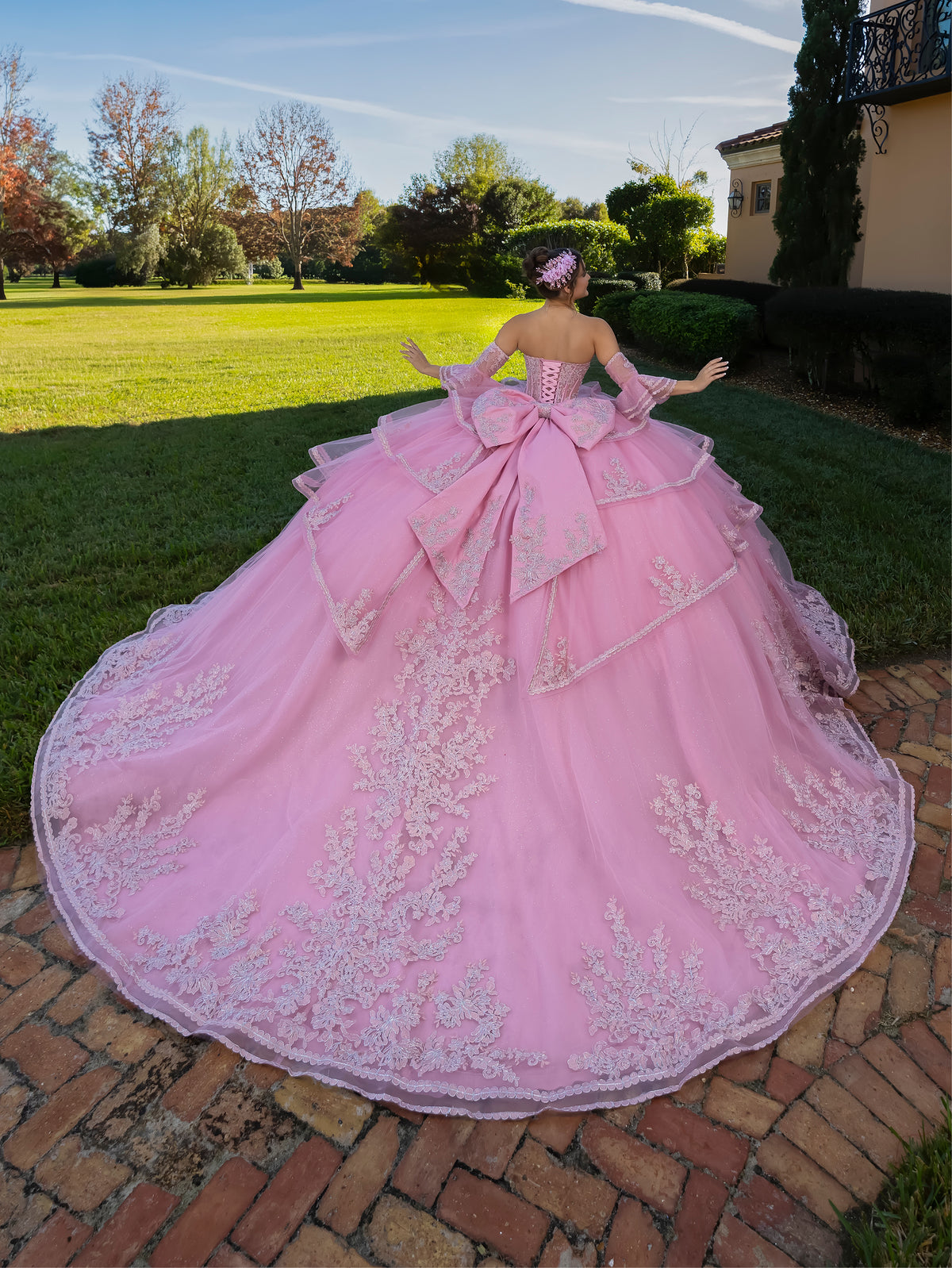 This ball gown features a fitted, embroidered bodice with off-the-shoulder sleeves. The full layered skirt includes cascading floral details and is finished with a dramatic bow and long, flowing train.