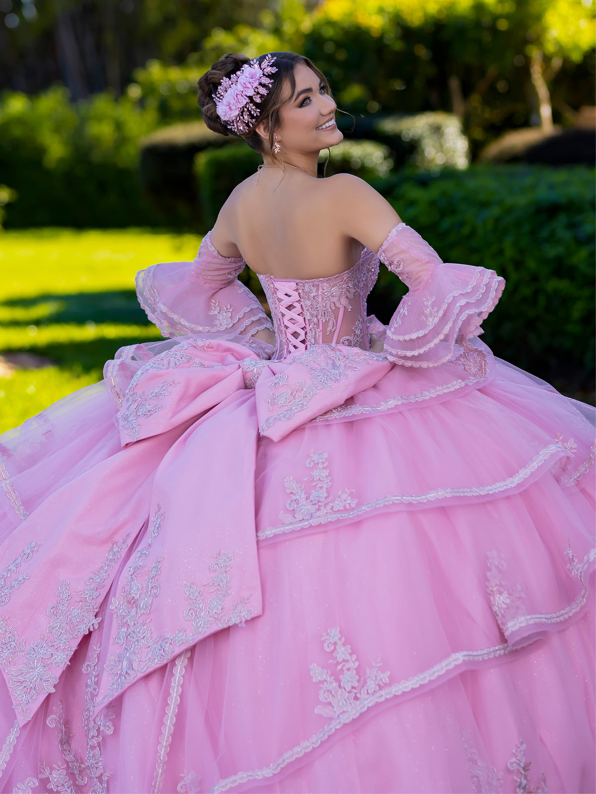 This ball gown features a fitted, embroidered bodice with off-the-shoulder sleeves. The full layered skirt includes cascading floral details and is finished with a dramatic bow and long, flowing train.