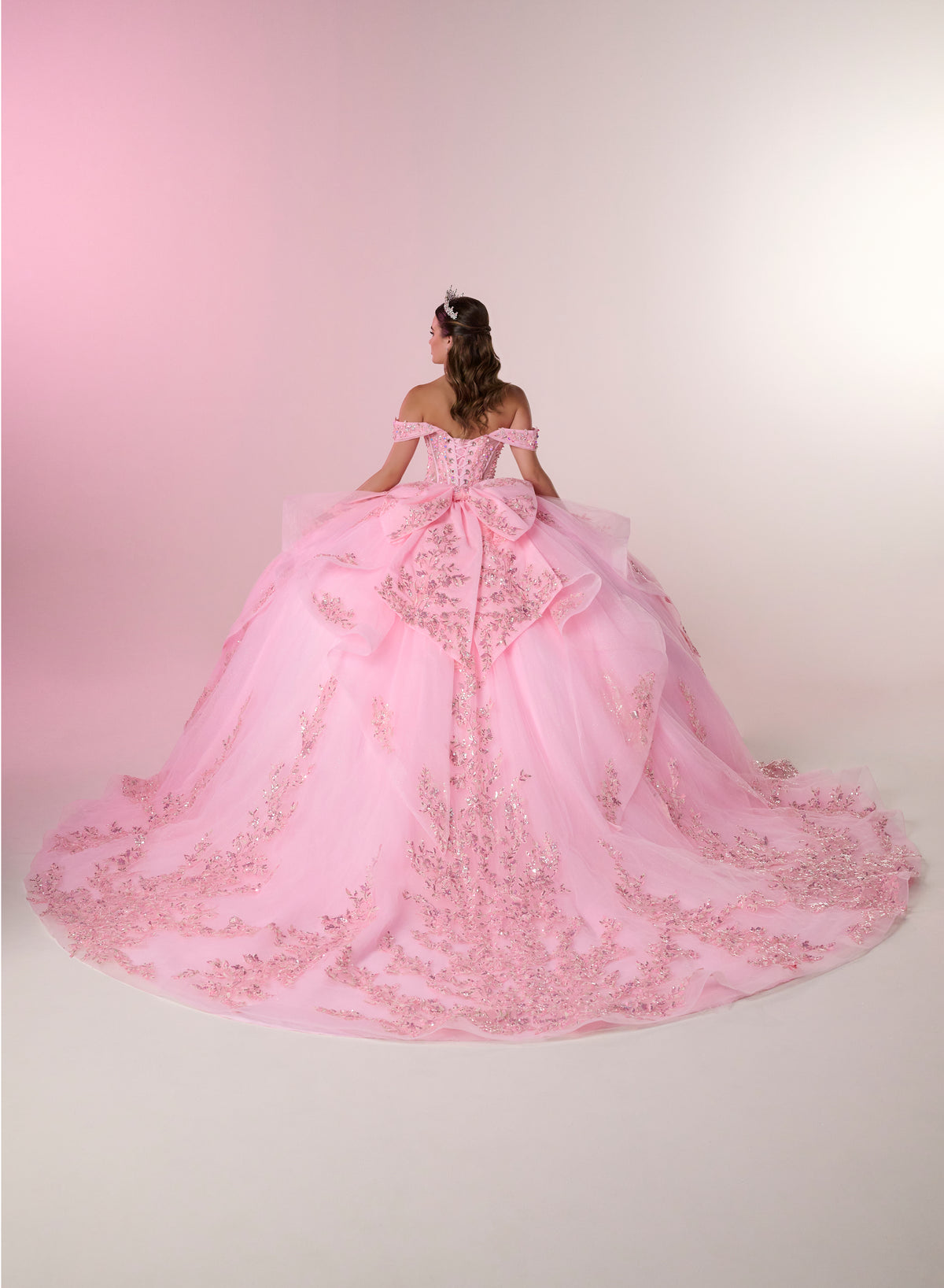 A woman viewed from behind wearing an elaborate off-the-shoulder pink ball gown with a large bow at the back and a long train covered in floral lace and beadwork, set against a soft pink studio background. / Stunning off-shoulder ball gown with shimmering embellished appliqués, a structured corset bodice, a full tulle skirt, and a dramatic oversized bow with a sweeping train.