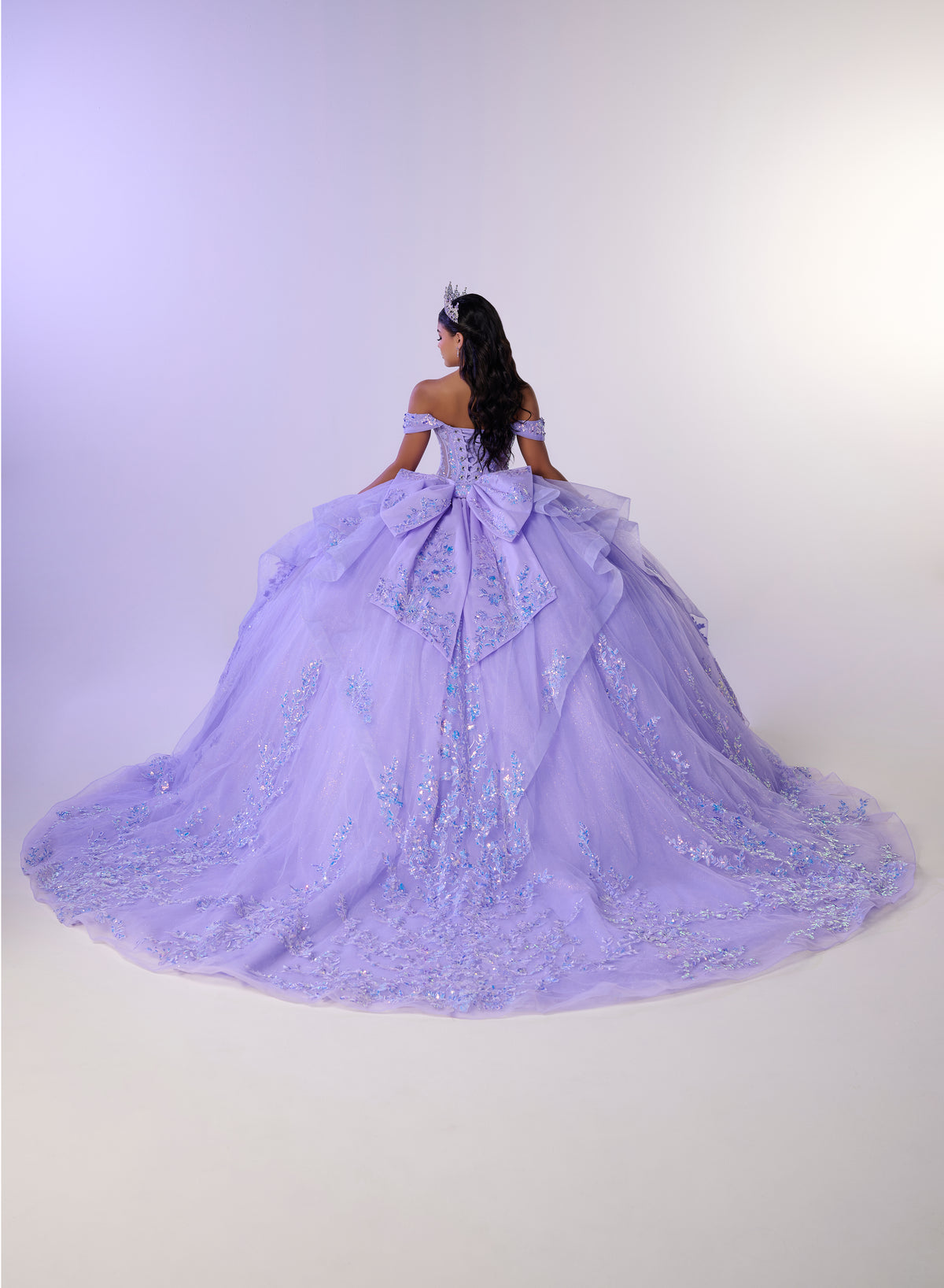 A woman viewed from behind wearing an elaborate off-the-shoulder lavender ball gown with a very full skirt, large bow at the back, and embroidered floral detailing, posed against a plain light studio background. / Stunning off-shoulder ball gown with shimmering embellished appliqués, a structured corset bodice, a full tulle skirt, and a dramatic oversized bow with a sweeping train.