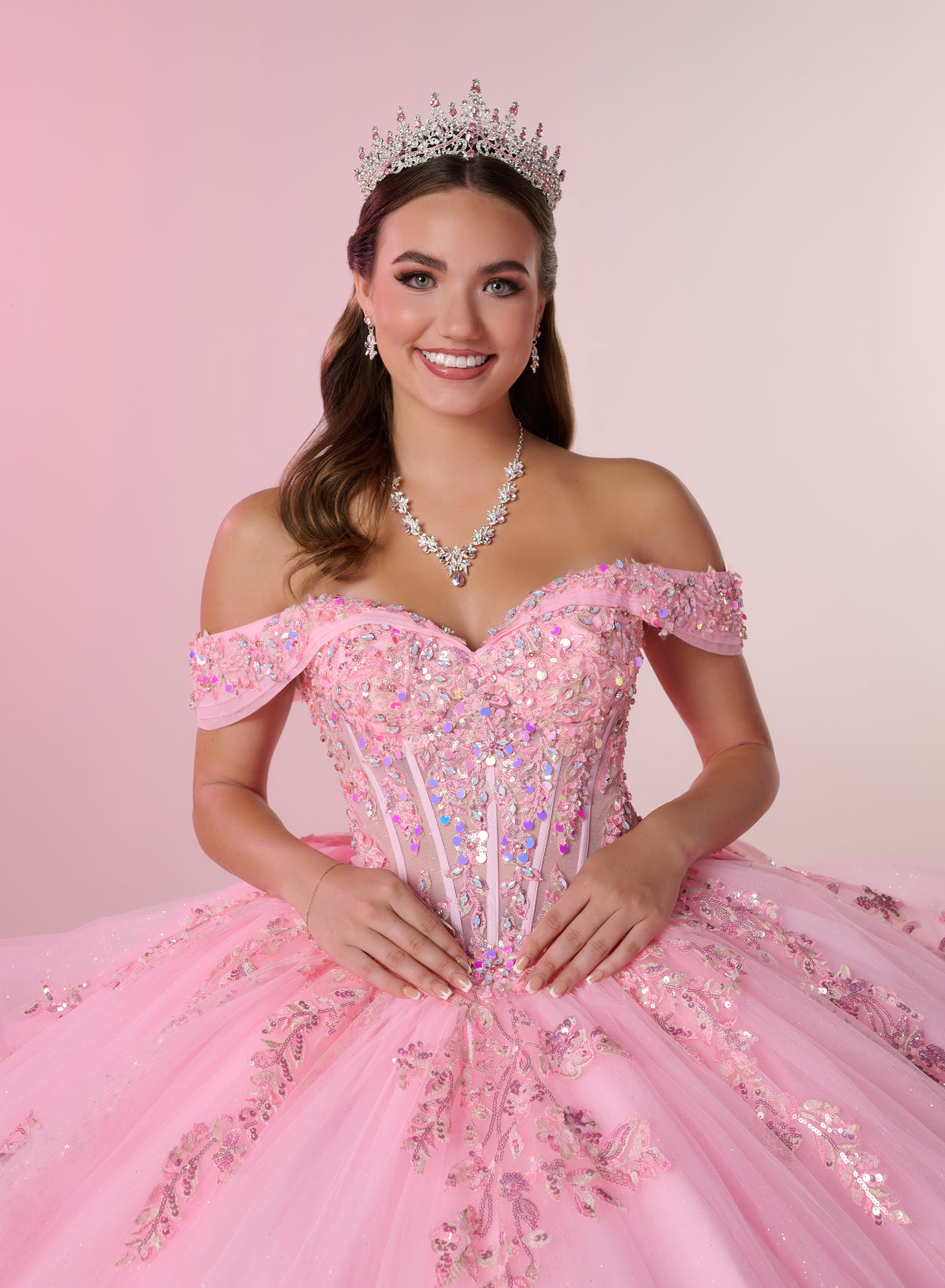 A smiling young woman wearing a sparkling off-the-shoulder pink ball gown with sequins and tulle, a silver tiara, and a matching silver necklace, posed against a soft pink studio background. / Stunning off-shoulder ball gown with shimmering embellished appliqués, a structured corset bodice, a full tulle skirt, and a dramatic oversized bow with a sweeping train.