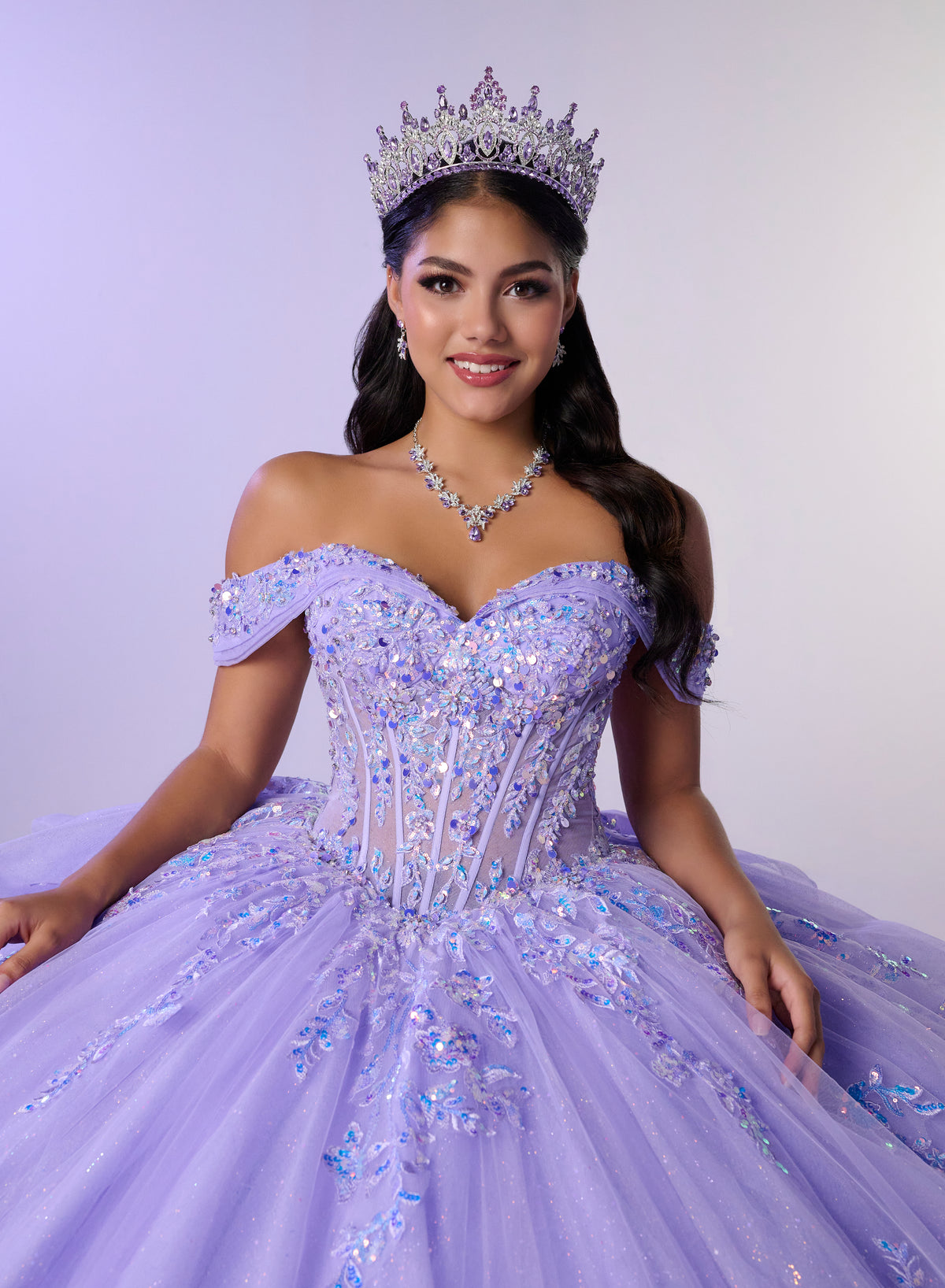 A young woman wearing a sparkling off-the-shoulder lavender quinceañera dress with blue sequin accents, a matching tiara, and a necklace, posed against a light lavender studio background. / Stunning off-shoulder ball gown with shimmering embellished appliqués, a structured corset bodice, a full tulle skirt, and a dramatic oversized bow with a sweeping train.