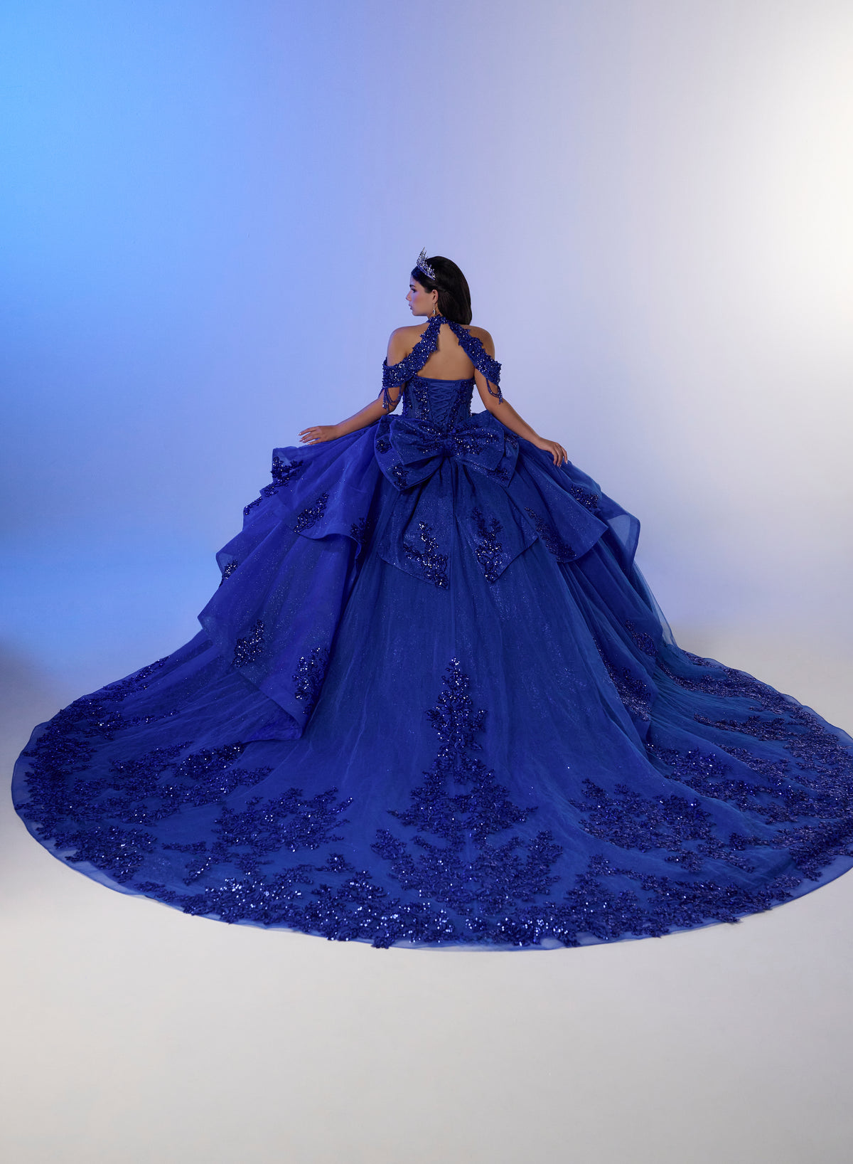 A woman wearing a voluminous royal blue ball gown with intricate lace and bead embellishments, photographed from behind against a blue-to-white studio backdrop. / Elegant ball gown featuring a beaded off-shoulder bodice, cascading ruffle details, intricate embellishments, and a dramatic oversized bow with a sweeping train.