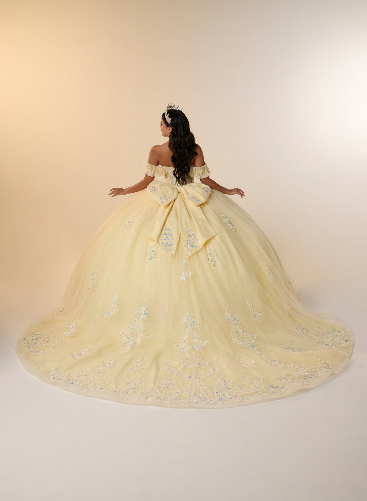A woman stands with her back to the camera wearing an off-the-shoulder pale yellow ball gown with a large bow at the back and light blue floral embroidery, set against a plain beige studio background. / Romantic off-shoulder ball gown with delicate embellished appliqués, a fitted bodice, a voluminous tulle skirt, and a statement bow at the back.